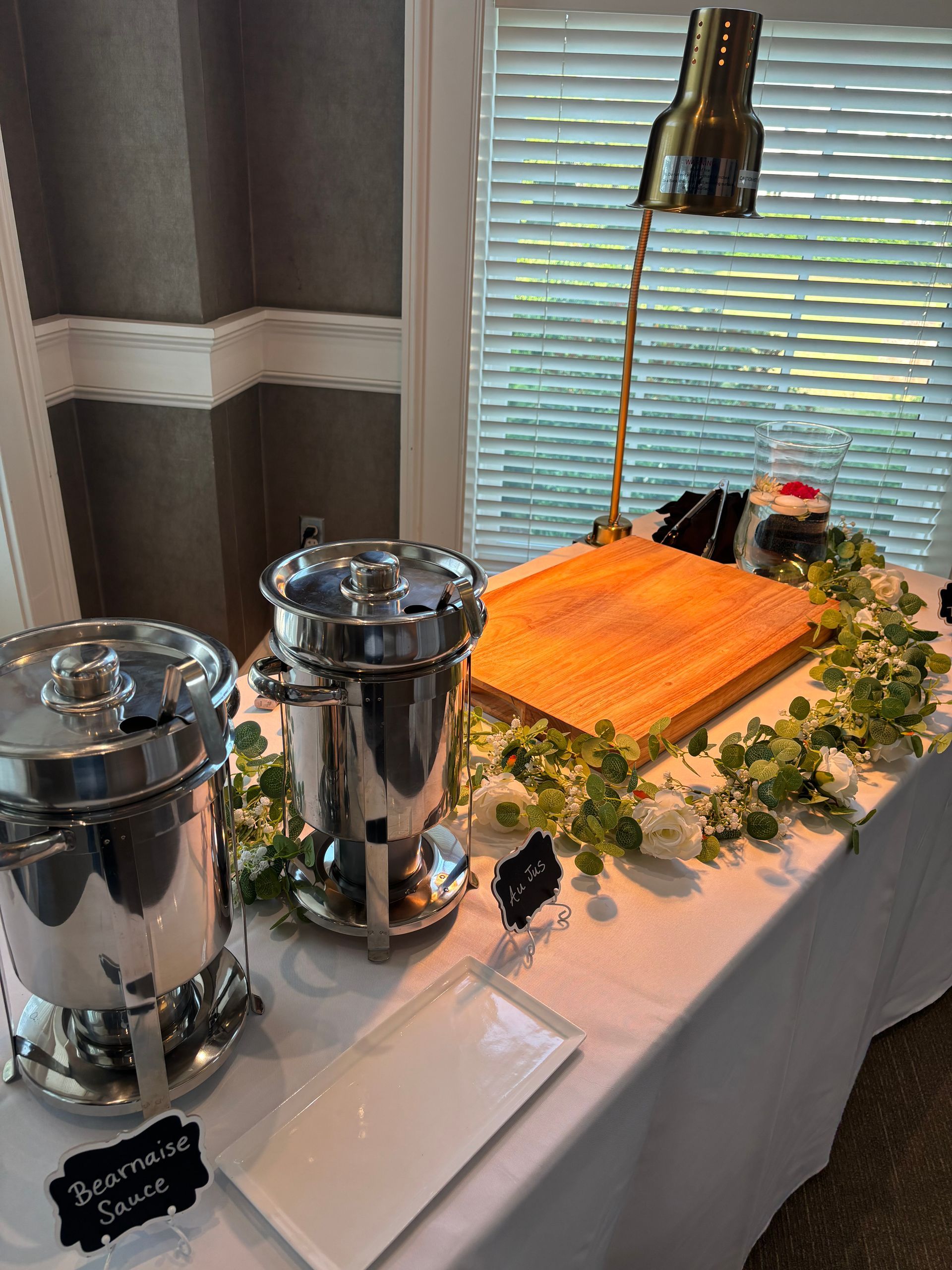 A buffet table with stainless steel food warmers, a wooden cutting board, and greenery, set against a window with white drapery.