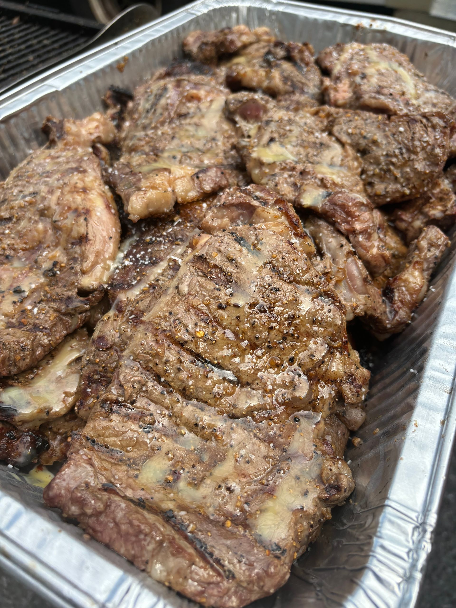 Grilled, seasoned ribs in a rectangular aluminum tray. The ribs are brown with a mix of spices and visible grill marks.