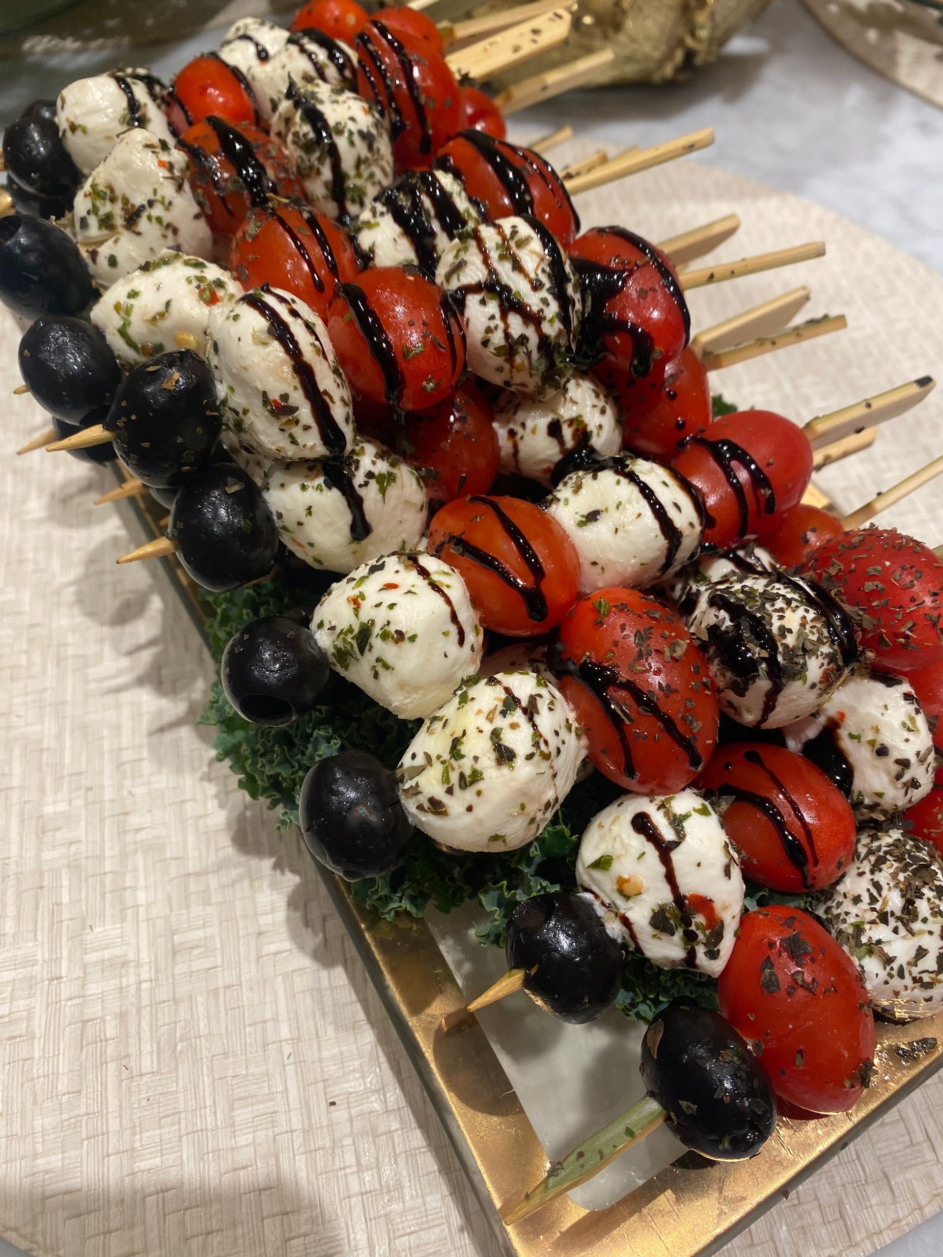 Caprese skewers with cherry tomatoes, mozzarella balls, and olives drizzled with balsamic glaze on a wooden tray.