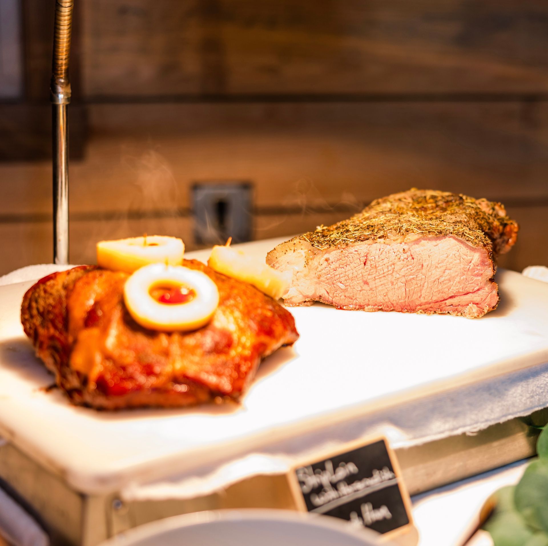 Roasted meats, including a piece with a crust of seasoning, on a serving board. Sliced potatoes and a sign with text are also visible.