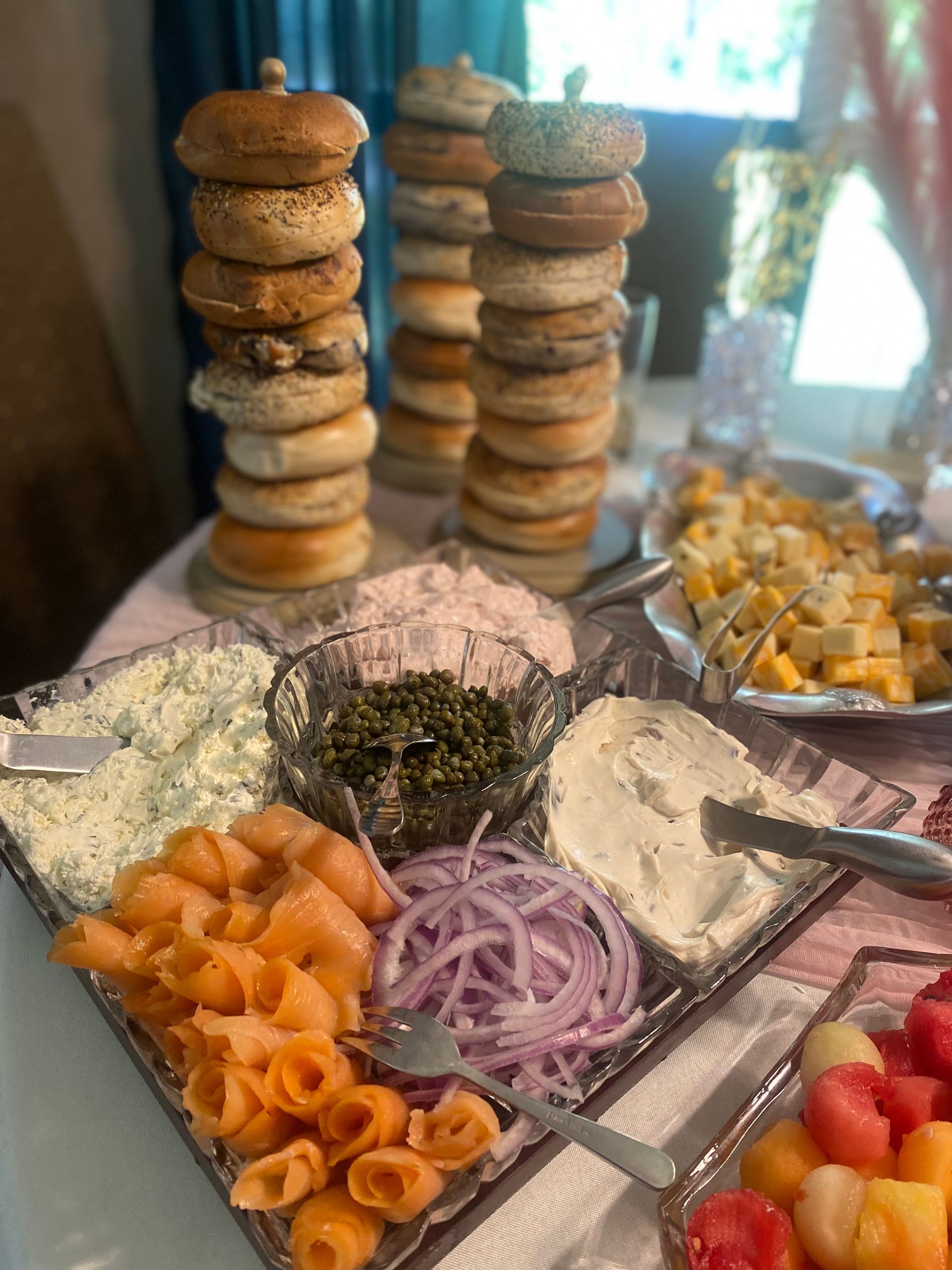 A brunch spread with bagel towers, smoked salmon, cream cheese, capers, and fruit on a table set for a meal.