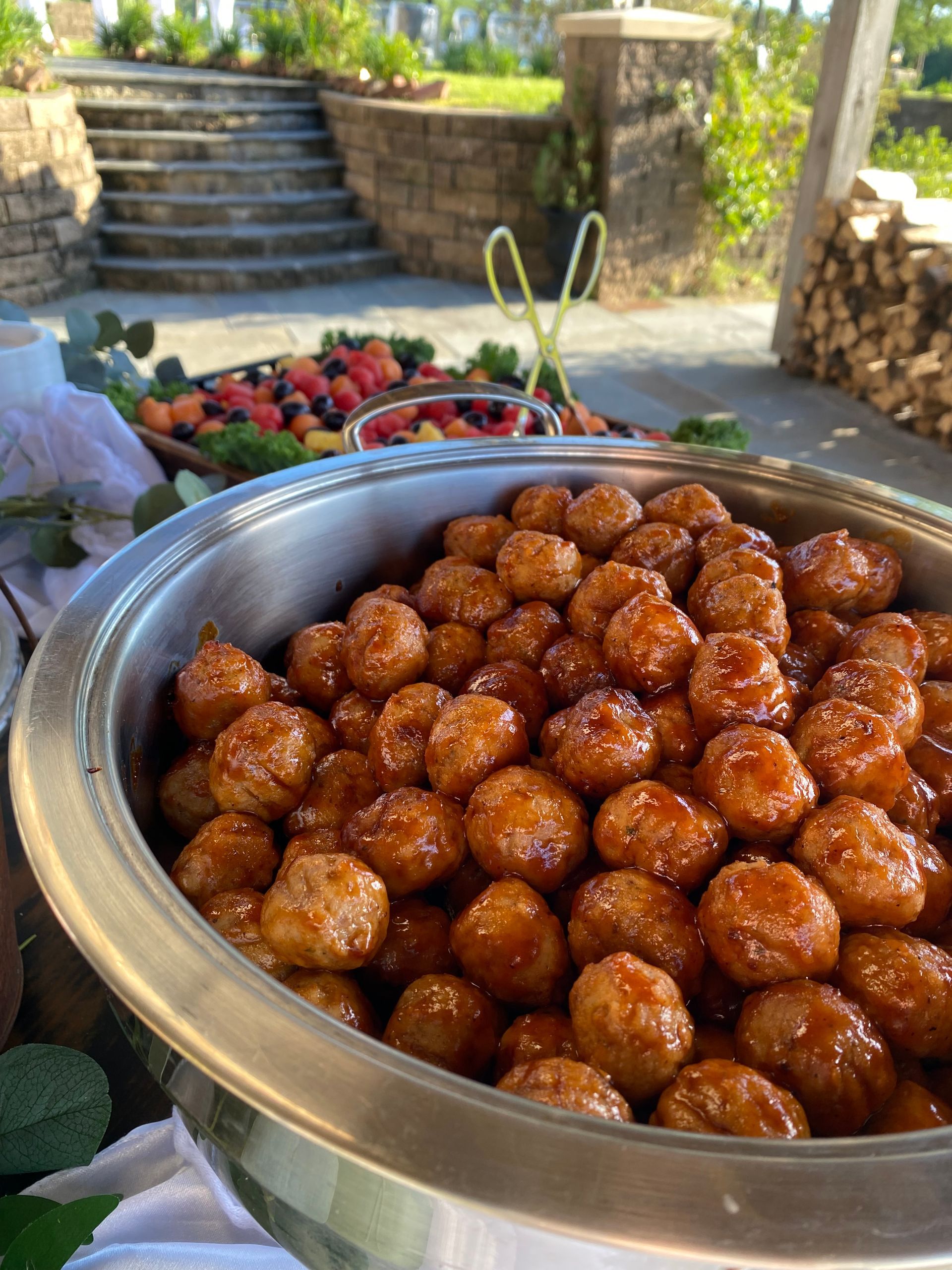 Large metal serving dish overflowing with glazed meatballs, with a blurred fruit platter and brick steps in the background.