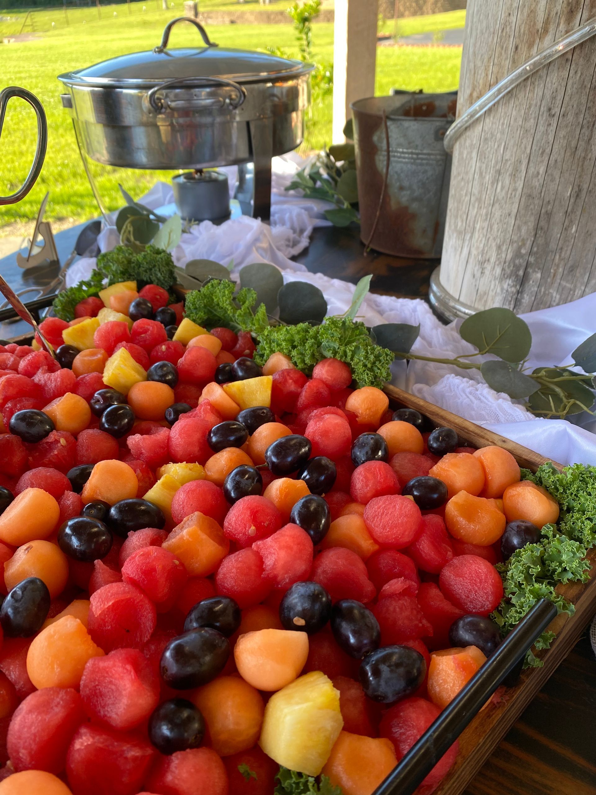 A wooden tray overflowing with colorful fruit, including watermelon, cantaloupe, pineapple, and grapes, arranged on a buffet table outdoors.