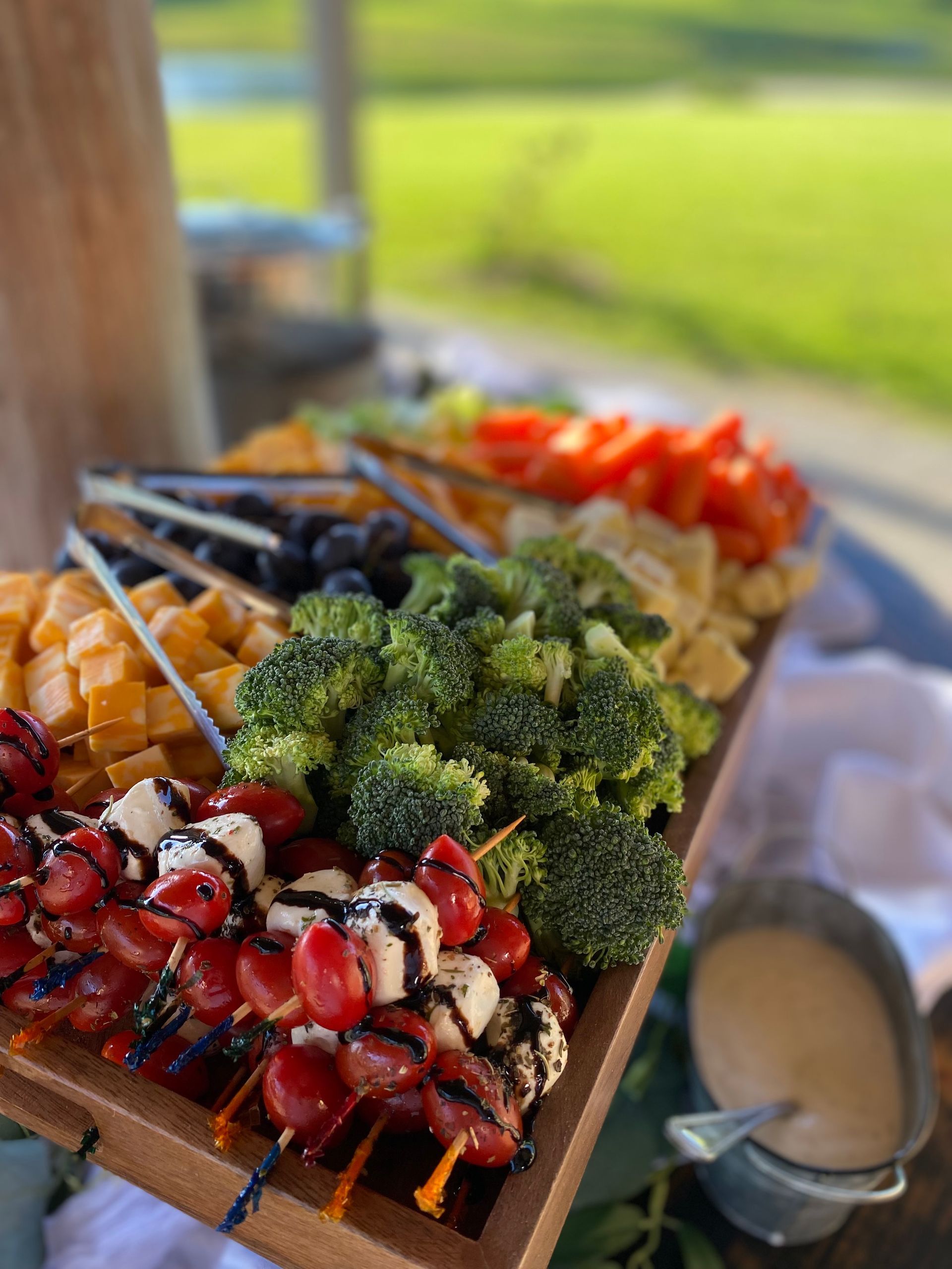 Wooden platter with colorful snacks: cheese cubes, broccoli, grapes, tomatoes, olives, and a dip. Set outdoors with a green field in the background.