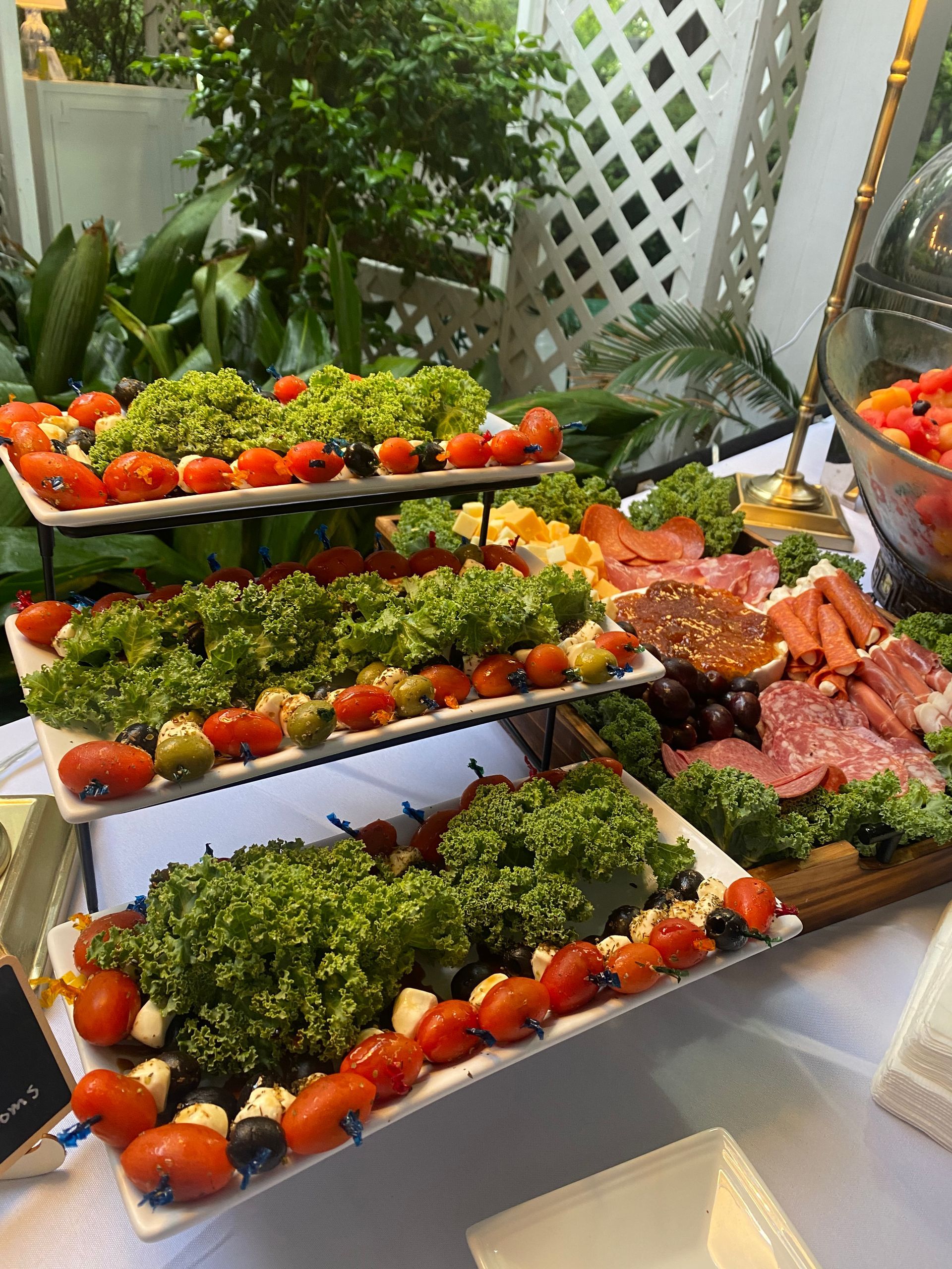 A buffet spread with layered platters of tomatoes, olives, and lettuce, alongside a charcuterie board and a bowl of tomatoes.