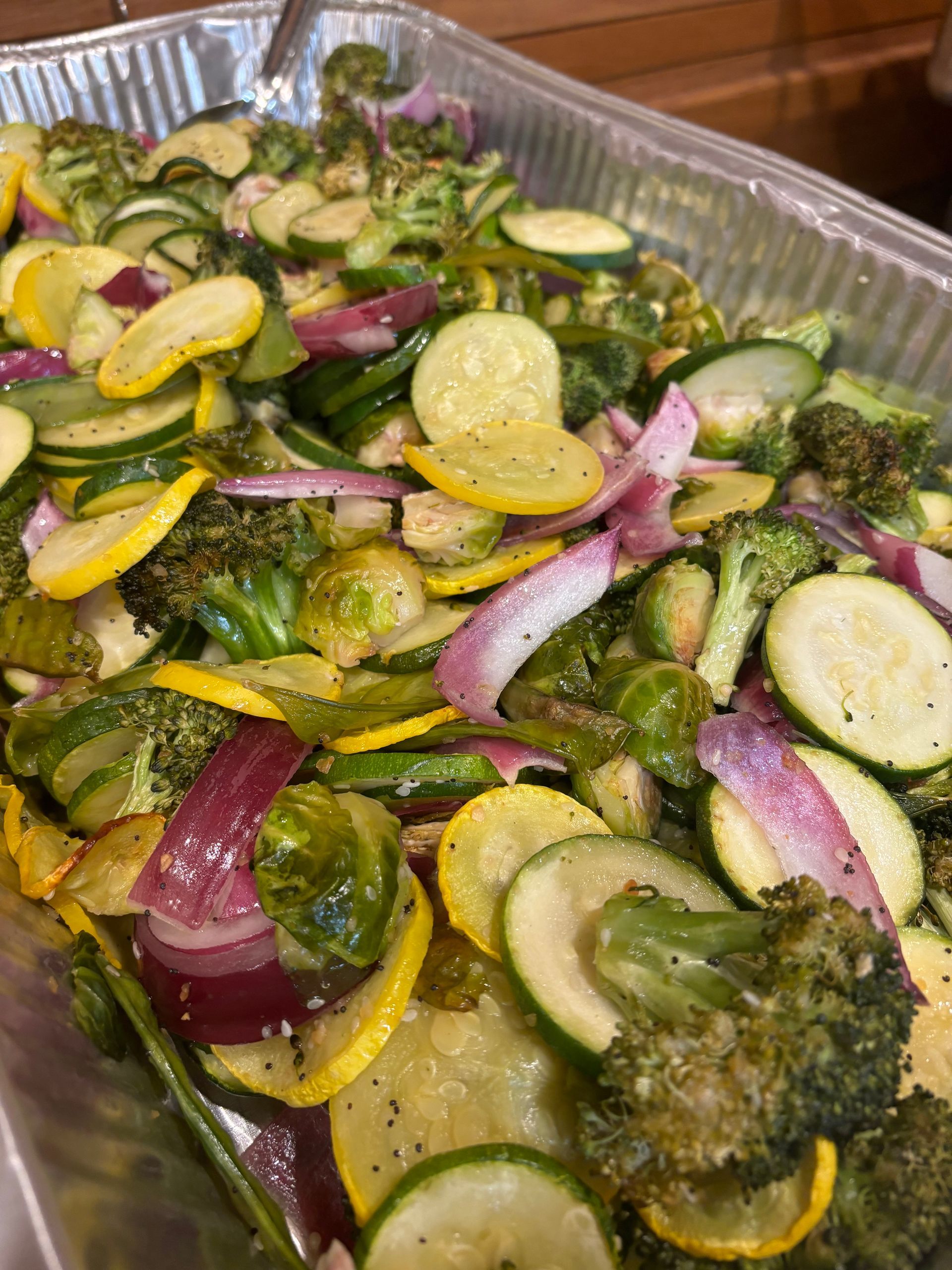 Roasted vegetables in a large metal tray, including zucchini, yellow squash, red onion, broccoli, and Brussels sprouts.