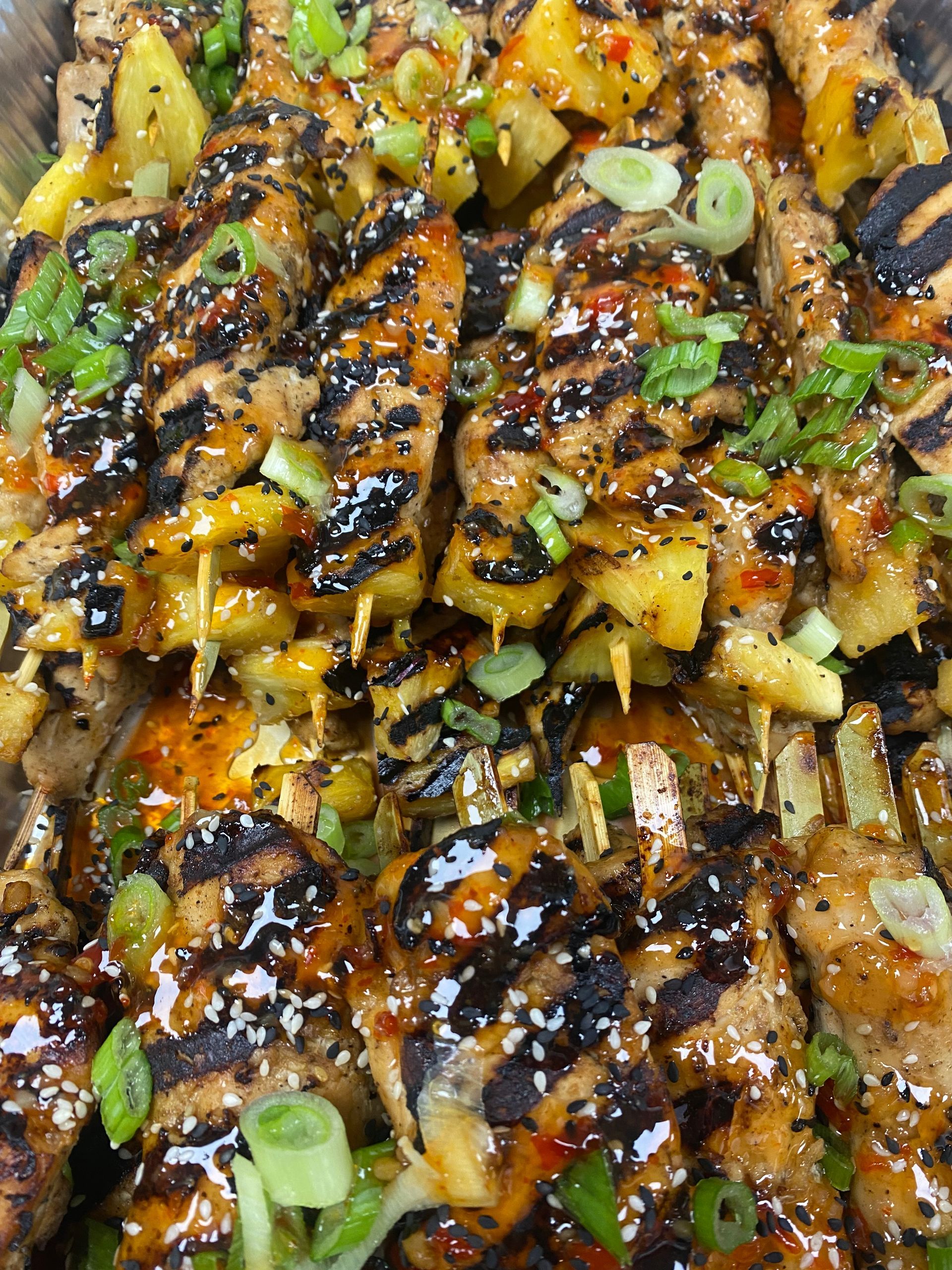 Grilled chicken and pineapple skewers drizzled with a sauce, garnished with sesame seeds and green onions.