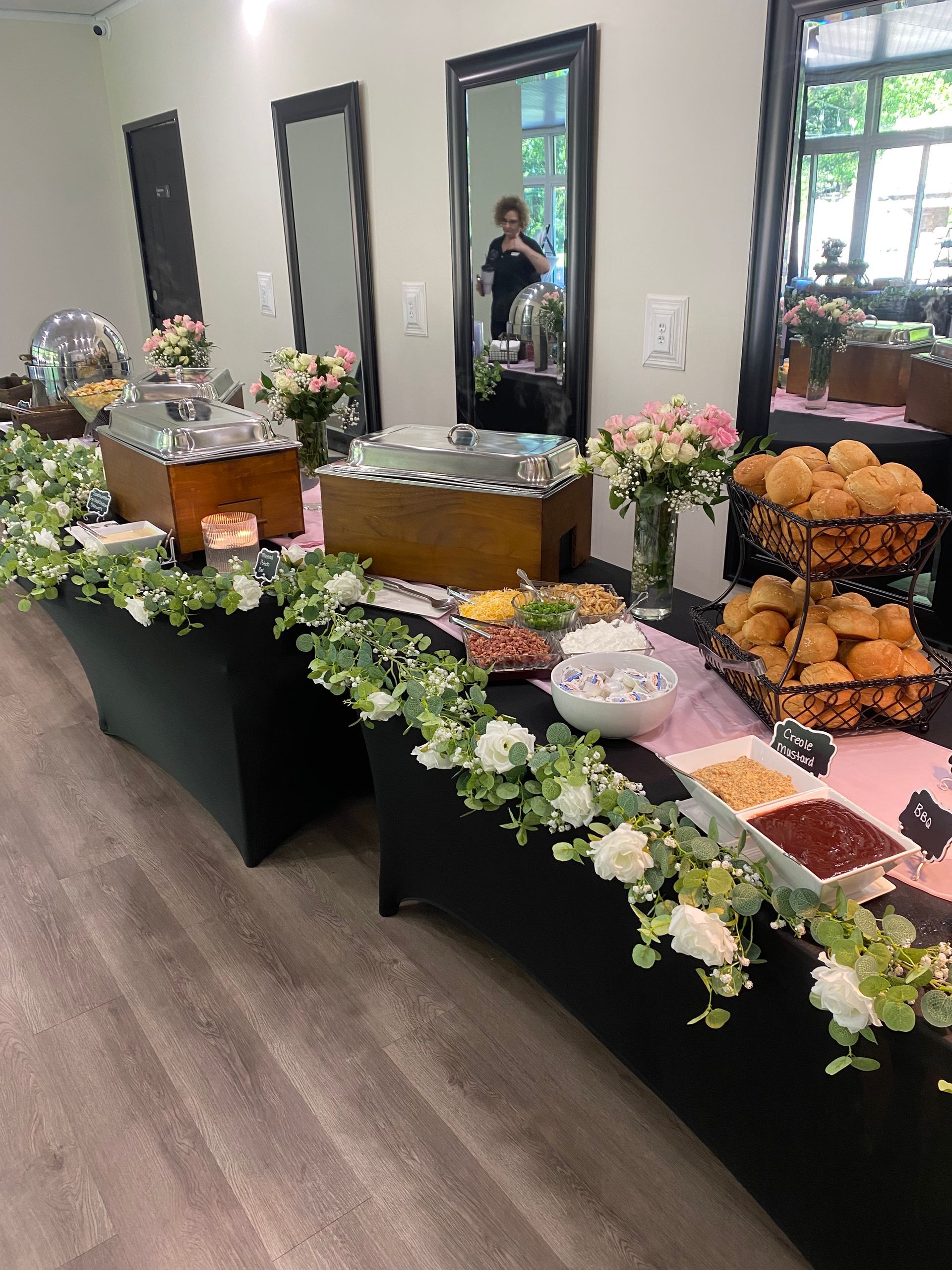 A food buffet table at an indoor event, featuring trays, bowls, and flowers. The table is black and has an array of food options.