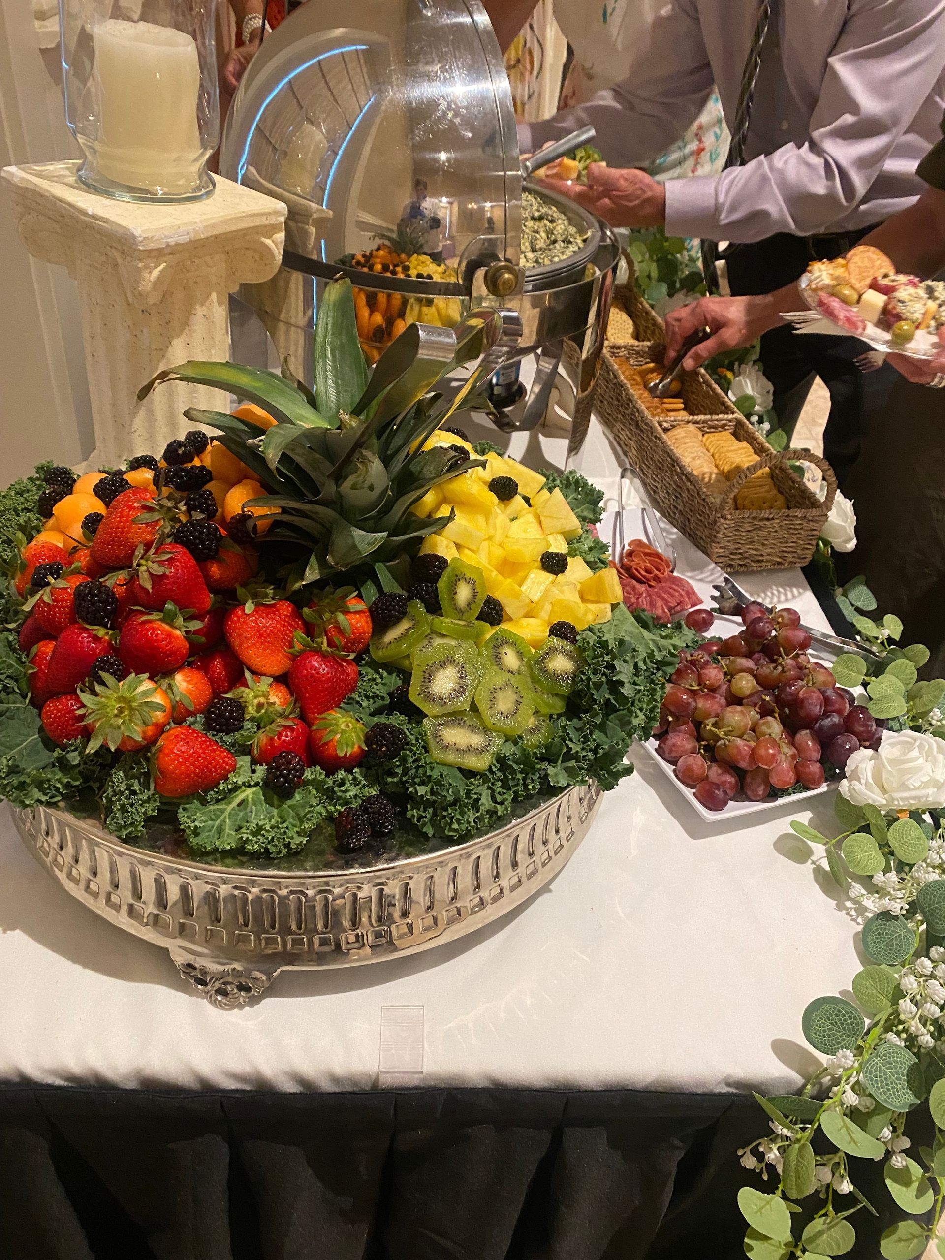 Fruit platter with strawberries, grapes, and other fruit. People are serving themselves from a buffet table.