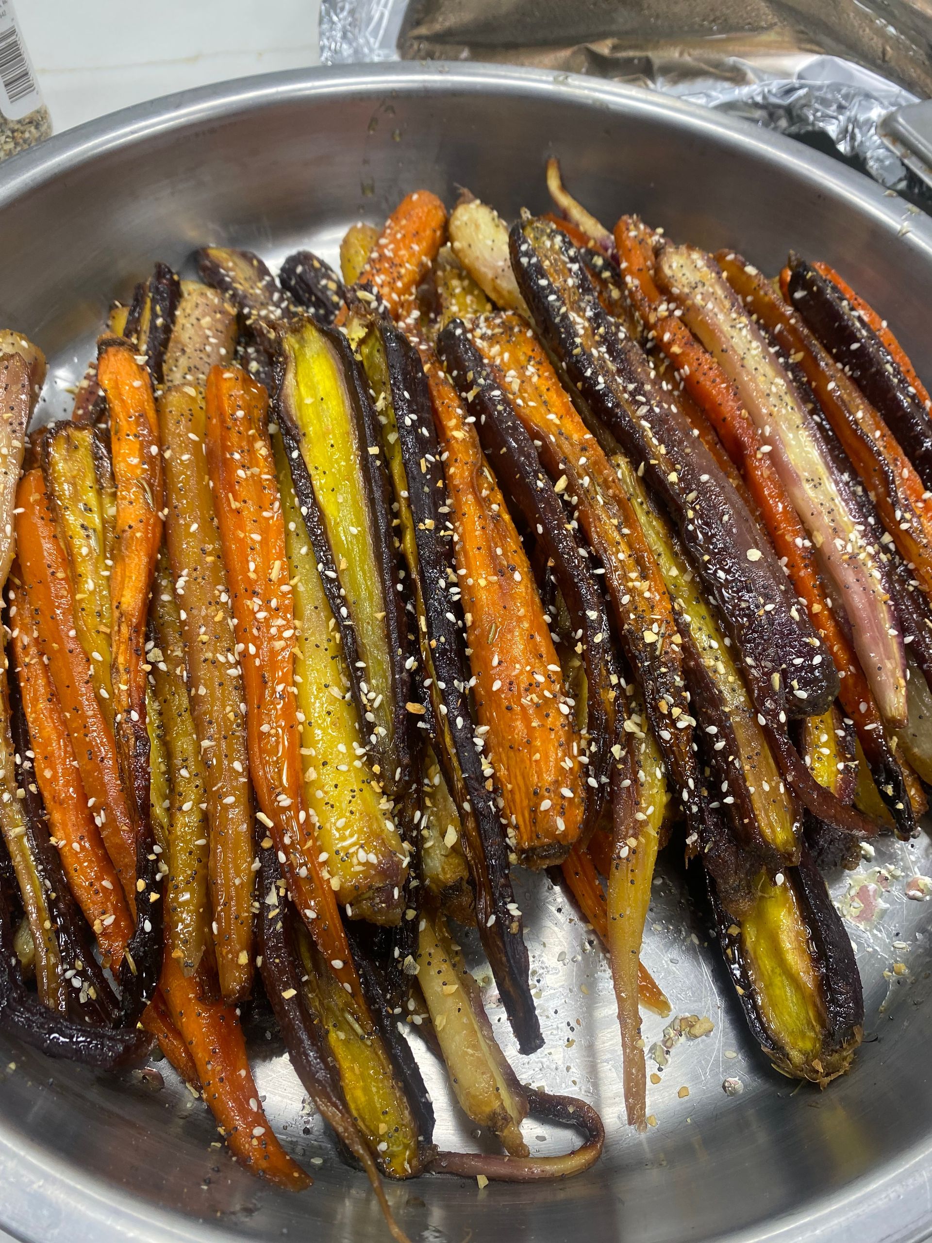 Roasted rainbow carrots in a metal bowl, sprinkled with sesame seeds. The carrots are orange, yellow, and purple.
