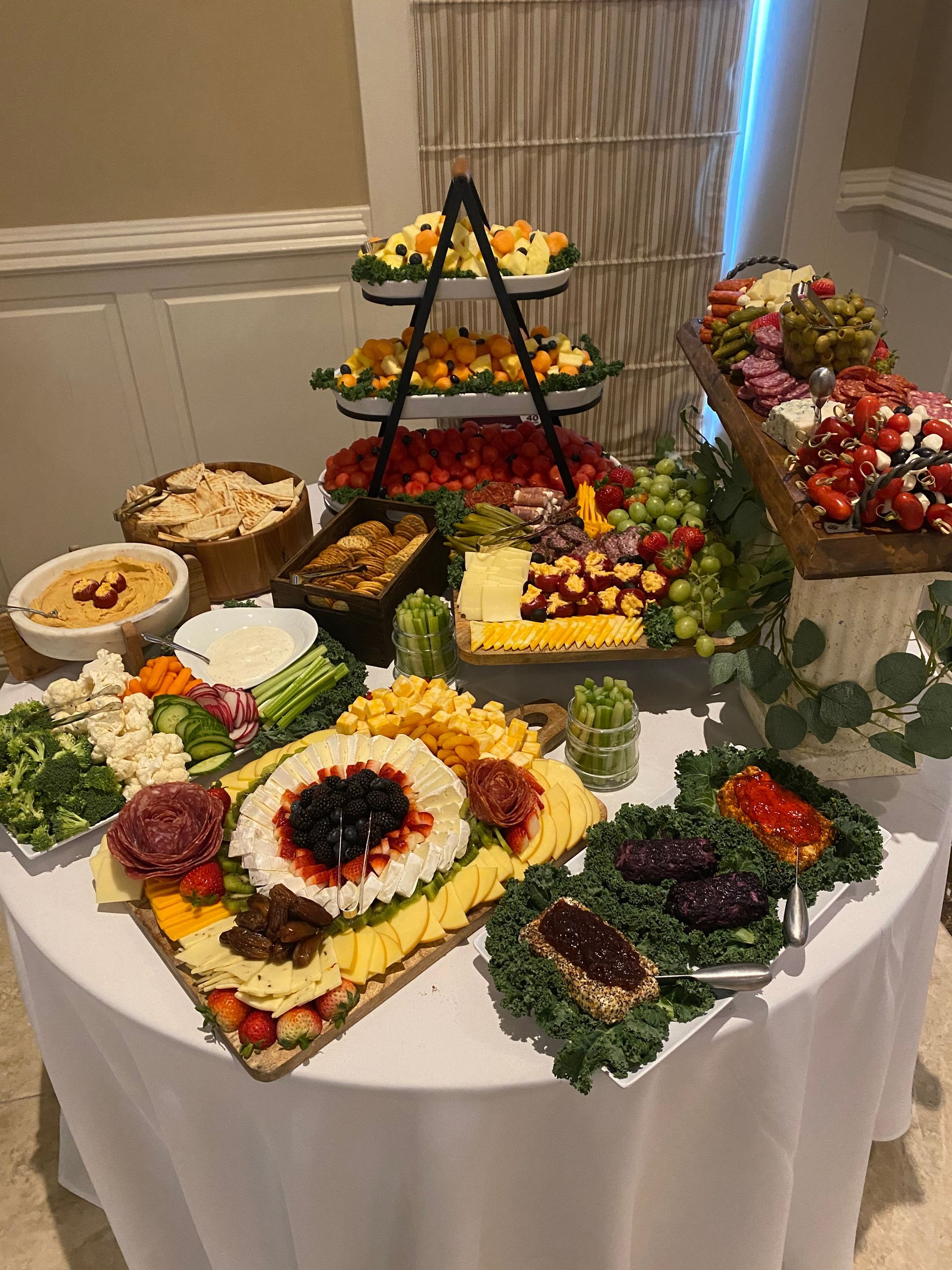 A round table with a white cloth is laden with charcuterie boards and platters featuring cheeses, fruits, vegetables, and dips, ready for a catered event.