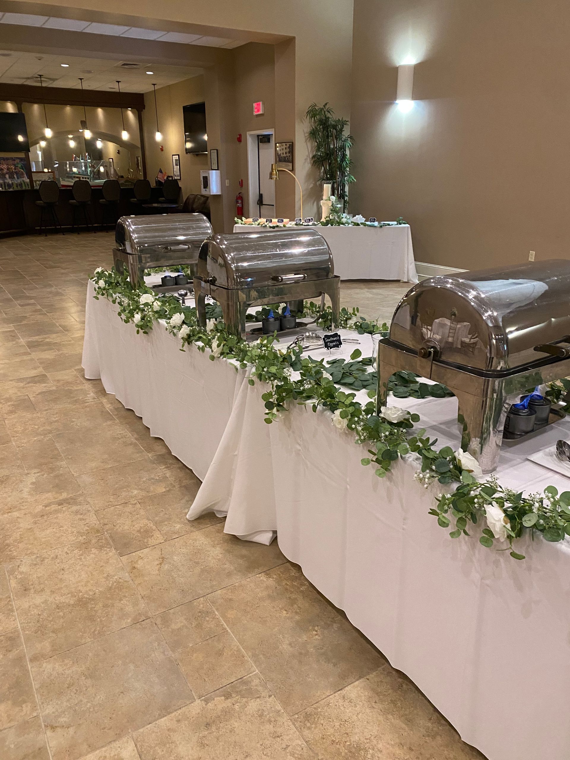 Buffet table with three silver food warmers, decorated with greenery. The setting is a beige-walled banquet hall.
