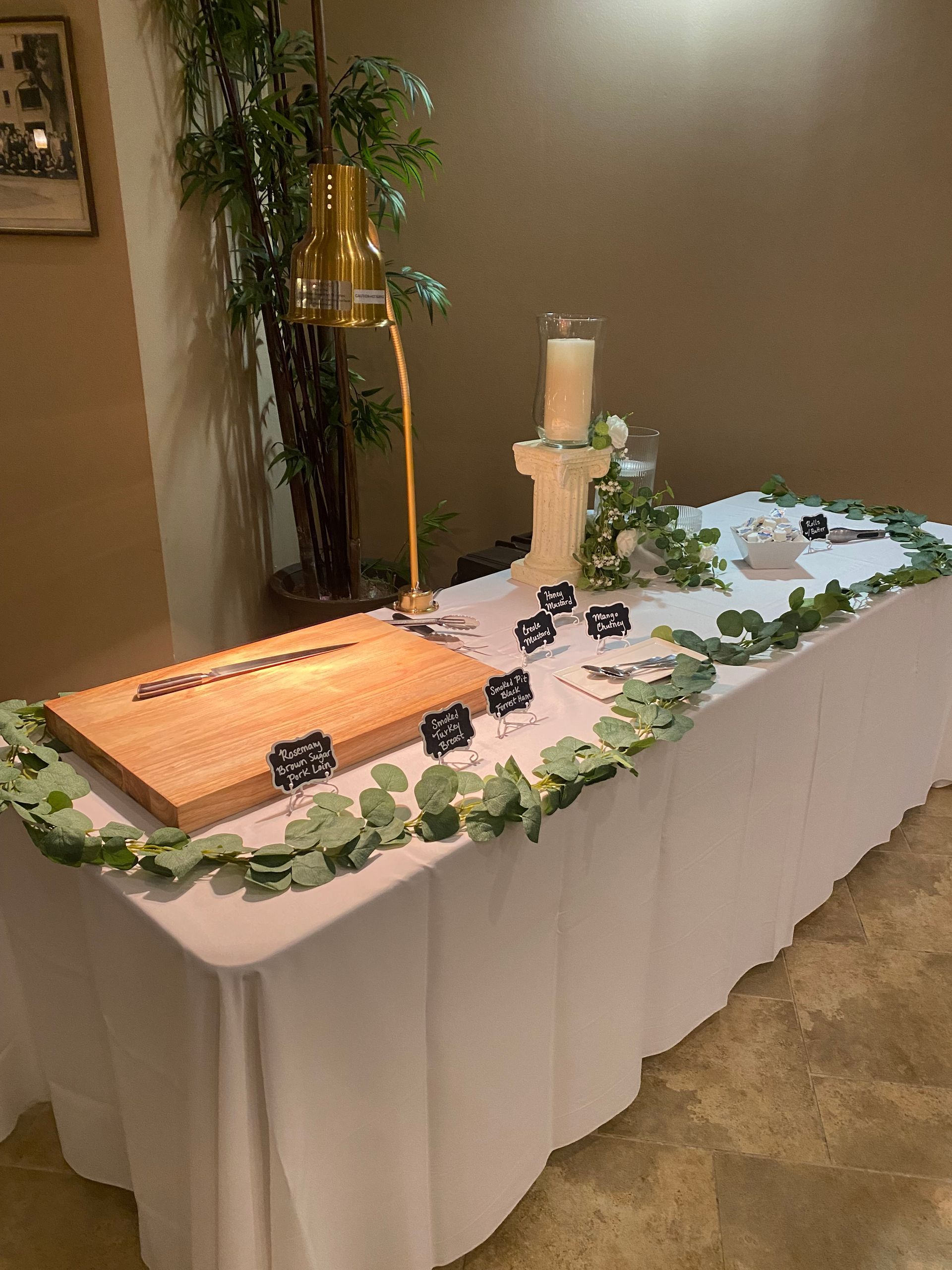A table draped in white with greenery, candles, and small chalkboards, likely for a wedding reception. A wooden cutting board is at the left.