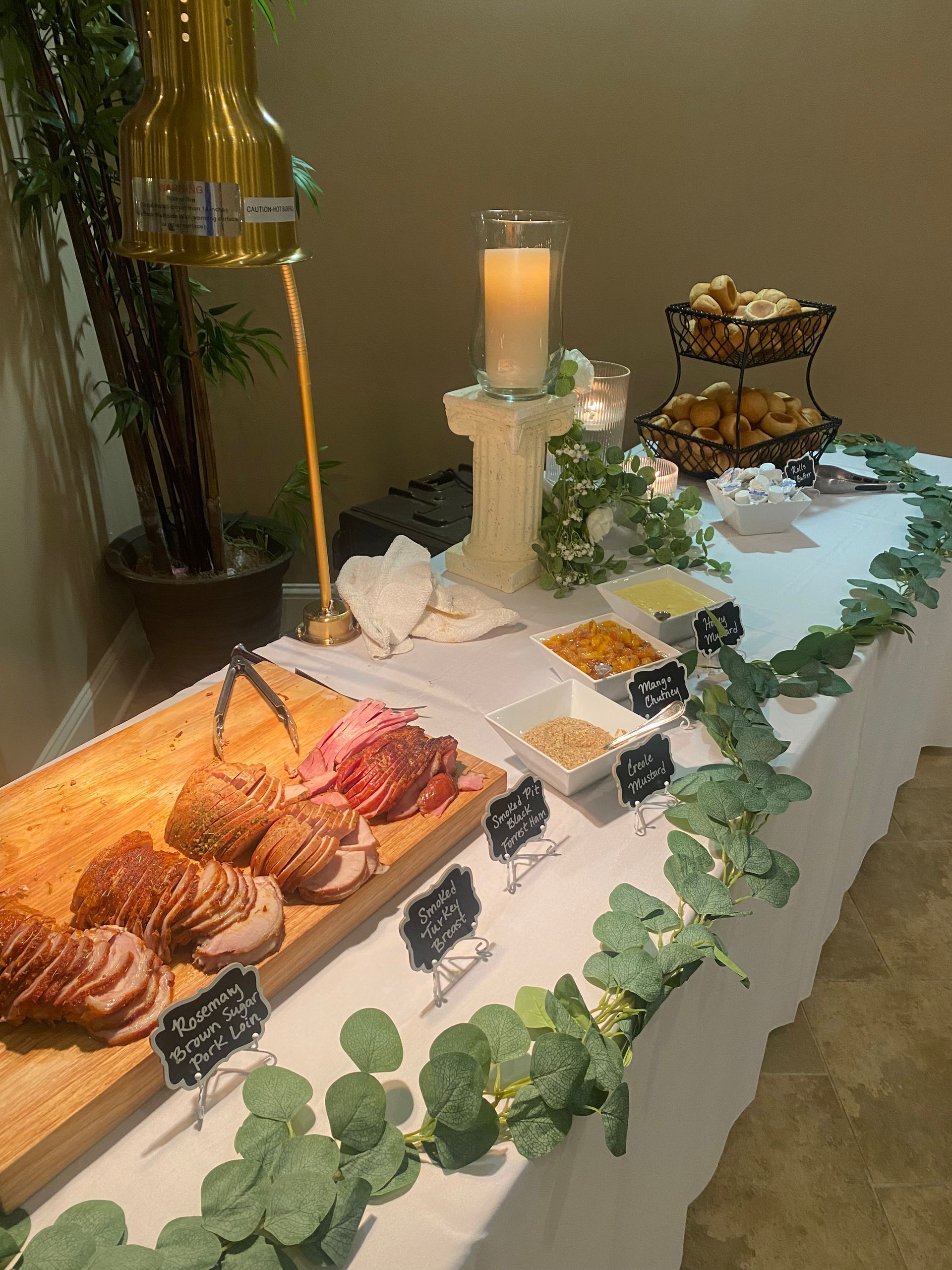 A buffet table with sliced meats, appetizers, and dipping sauces arranged on a white tablecloth, decorated with greenery.