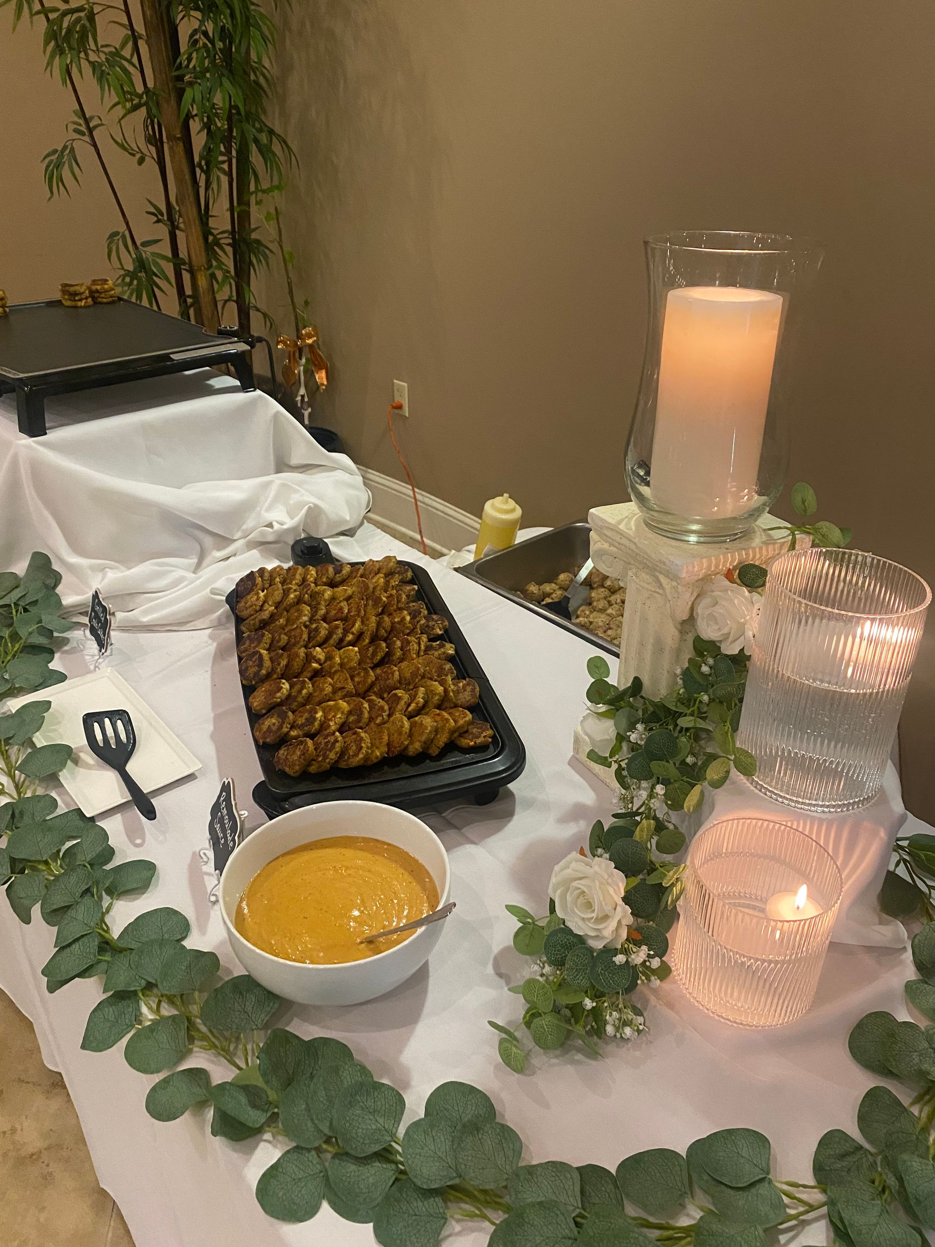Buffet table with food, decorations, and a lit candle. Features include a tray of food, a bowl of dip, and greenery.