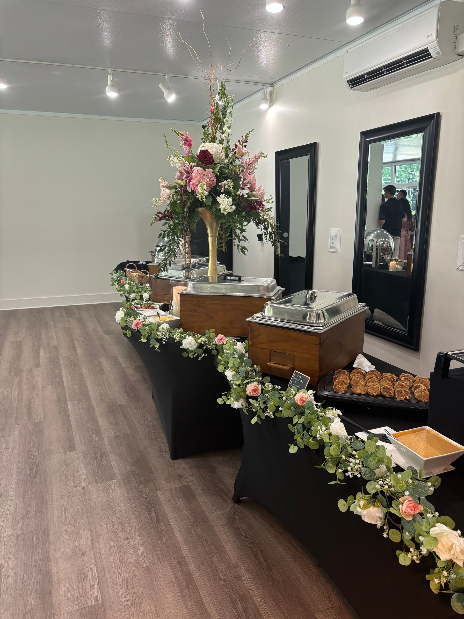 Buffet table at an event with floral arrangements and food displays. Black tablecloths, light wood flooring, and a white wall.