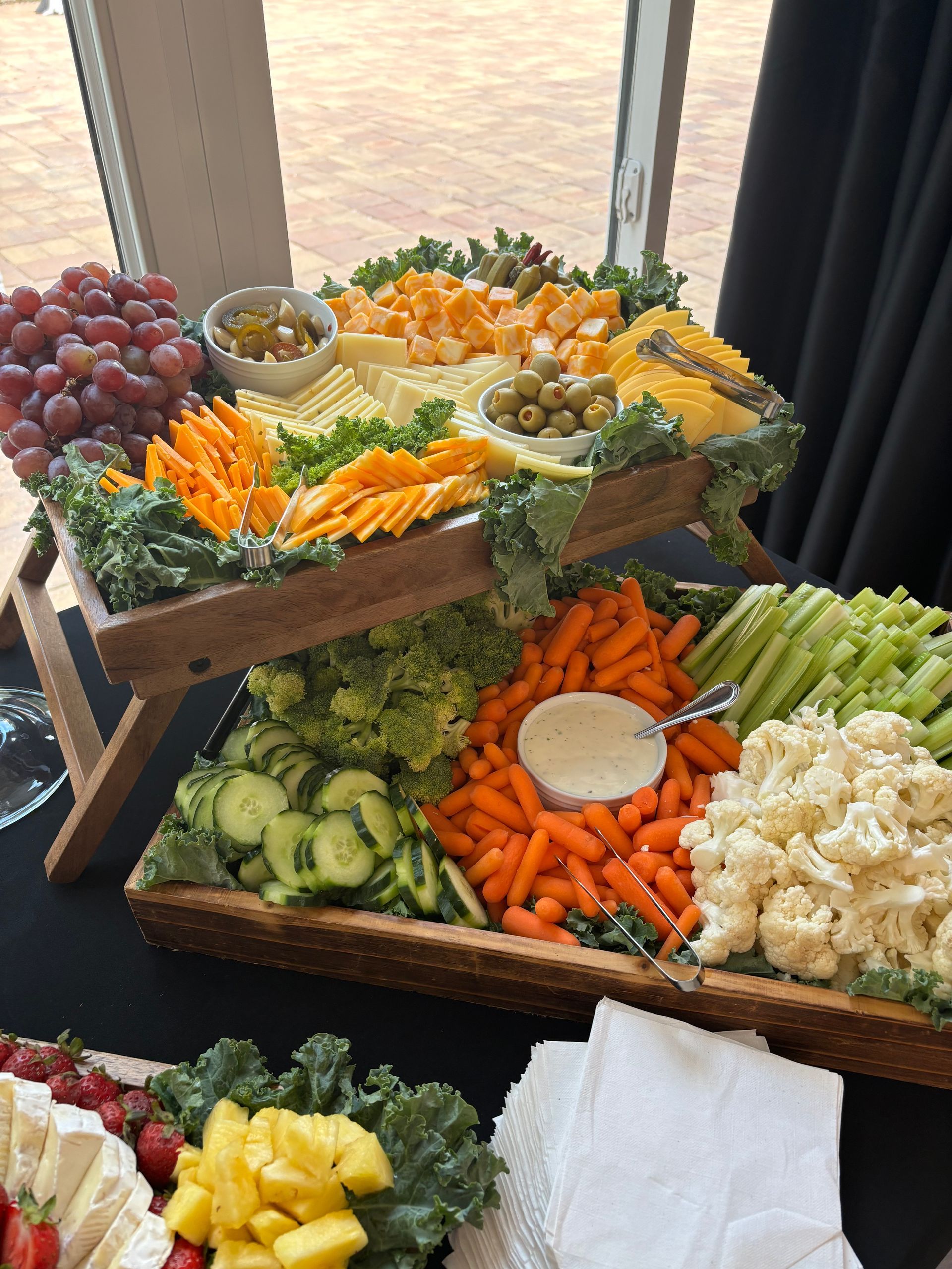 A tiered charcuterie board with cheese, vegetables, grapes, and dipping sauce, set on a table near a window.