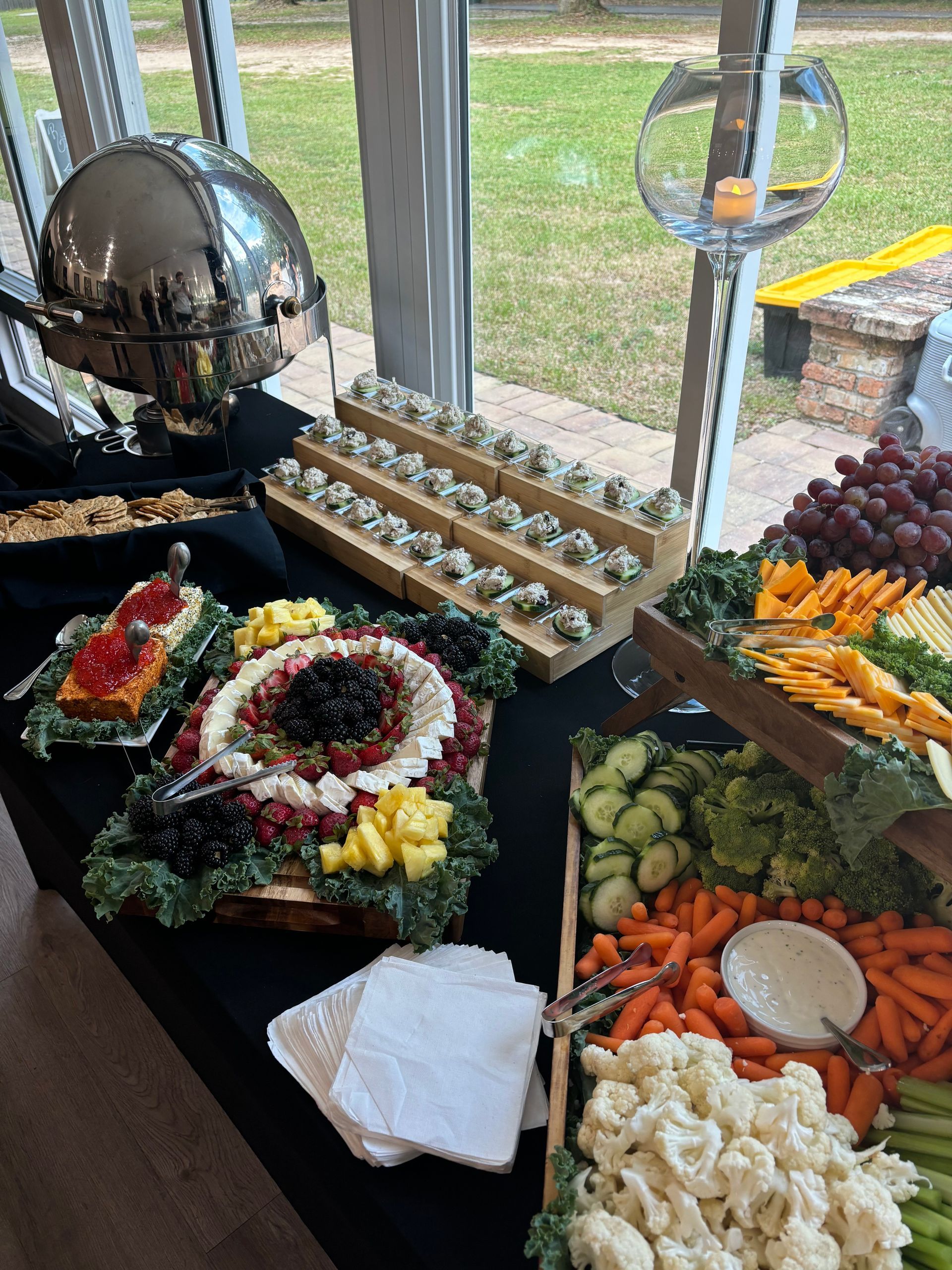 Buffet table with various foods like fruits, vegetables, and appetizers. Window backdrop.