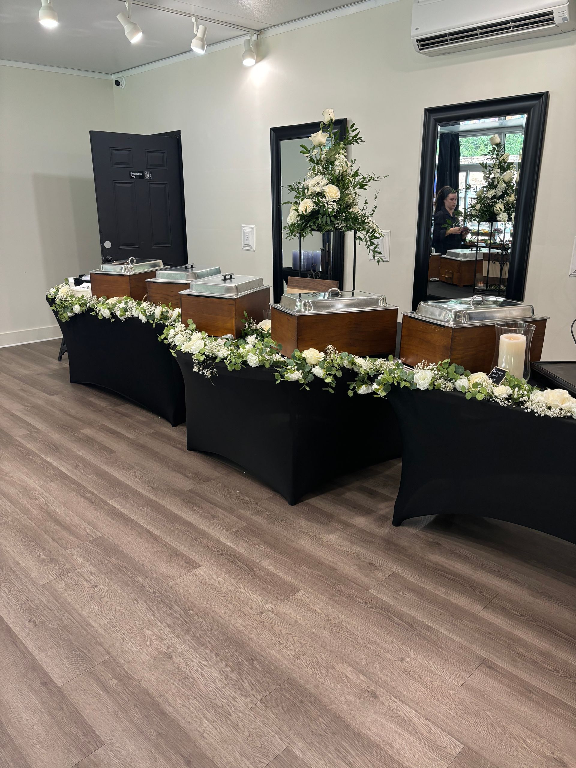 Buffet tables draped in black, holding food and decorated with white flowers, set in a room with light wood floors and large mirrors.