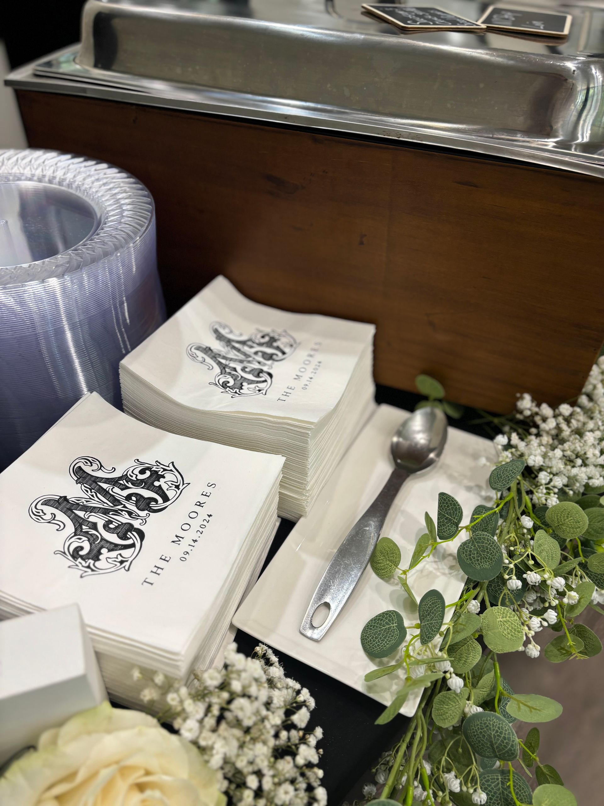 Napkins with a crest and text, a spoon, and floral decor near a buffet warmer.