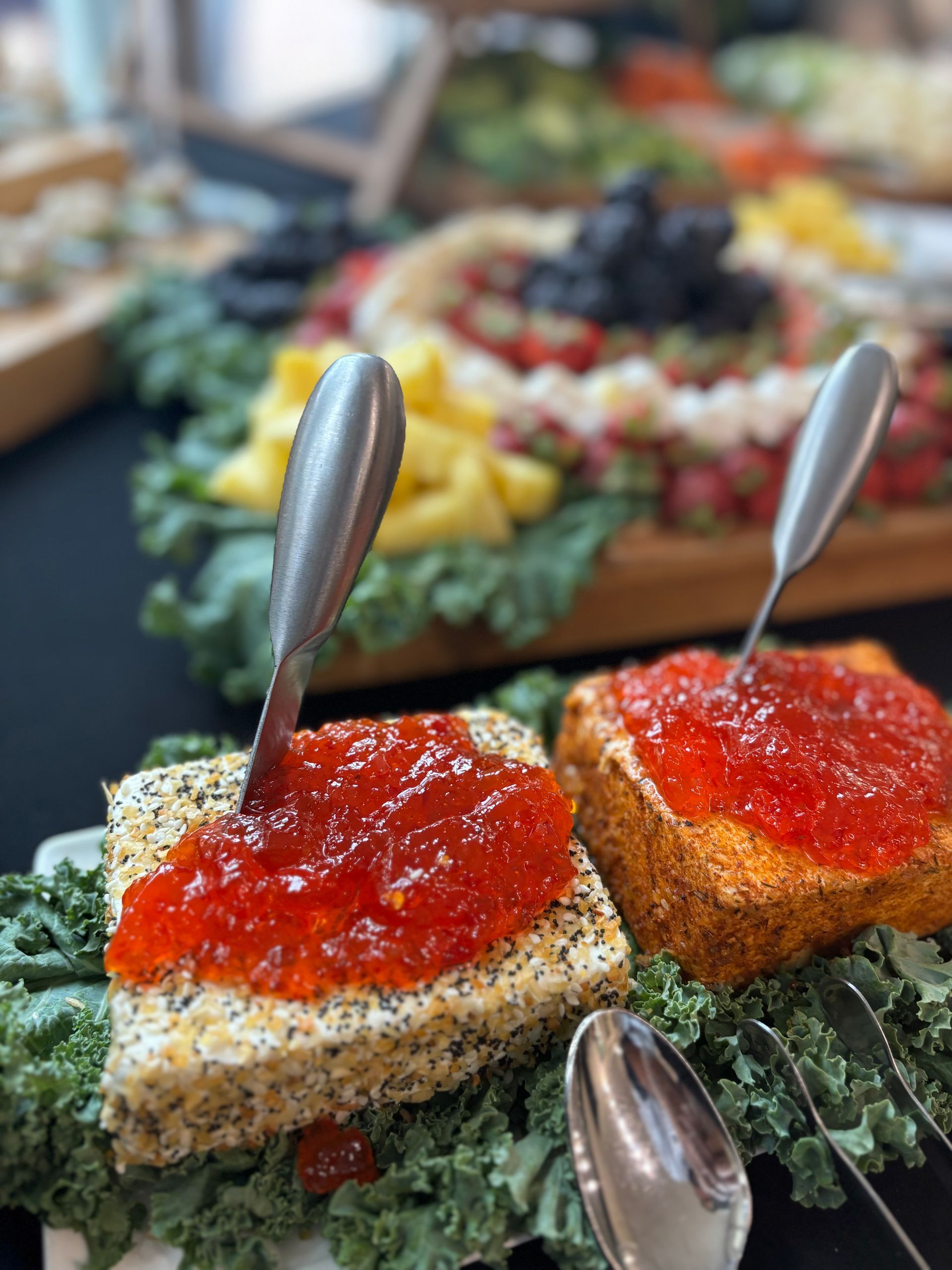 Two cheese appetizers topped with red jam, each with a spoon, sit on a bed of kale. Other appetizers are blurred in the background.