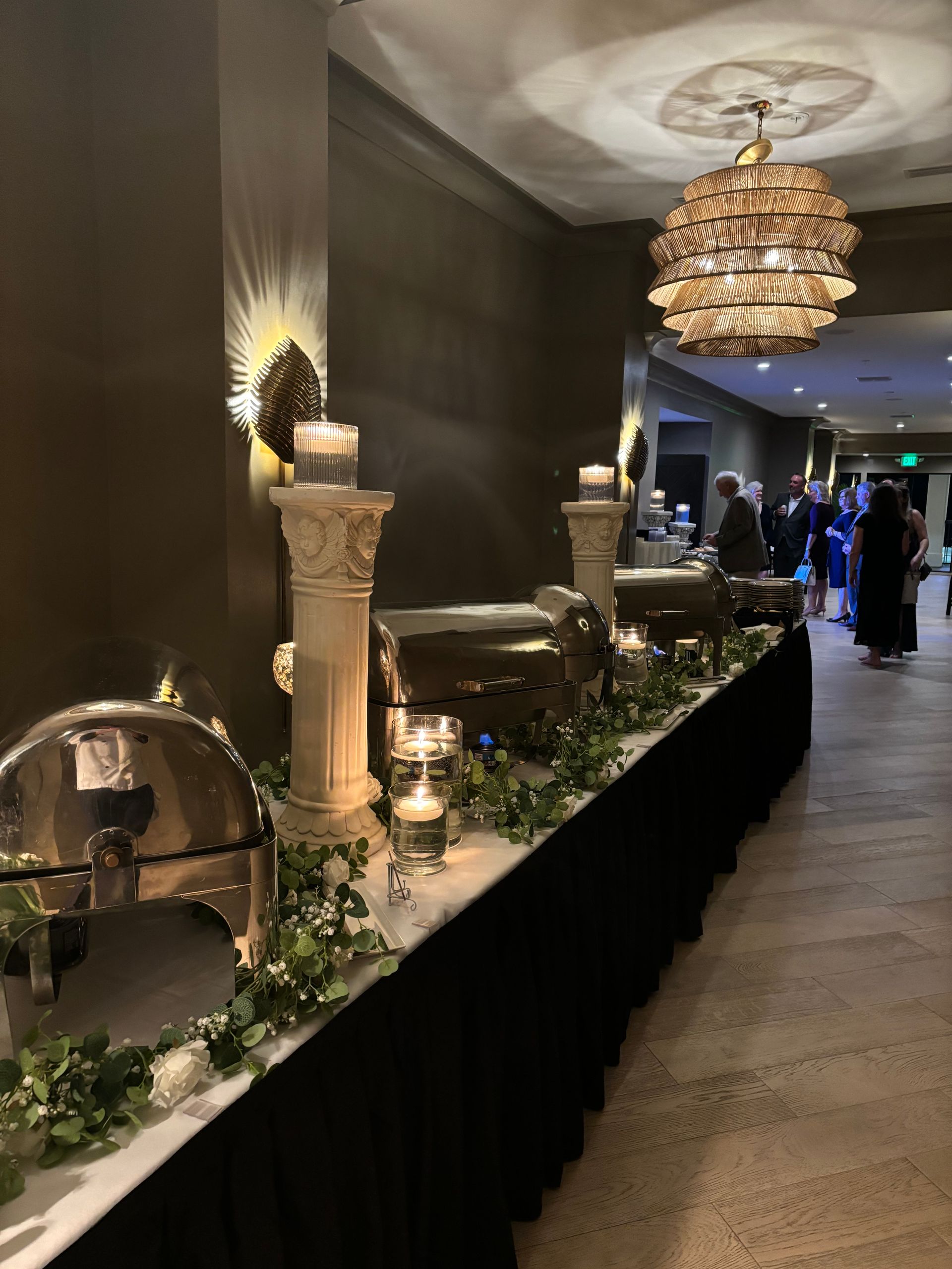 Buffet table at an event. Long, black-skirted table with chafing dishes, pillars, candles, and greenery. People in formal attire are visible in the background.