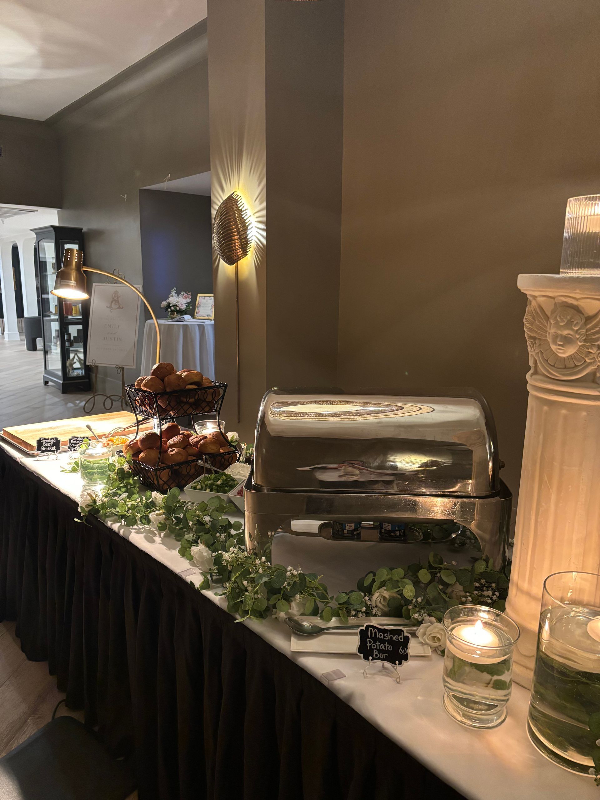 Buffet table with food, chafing dishes, and decorations. The table is set against a wall with sconces and a column.