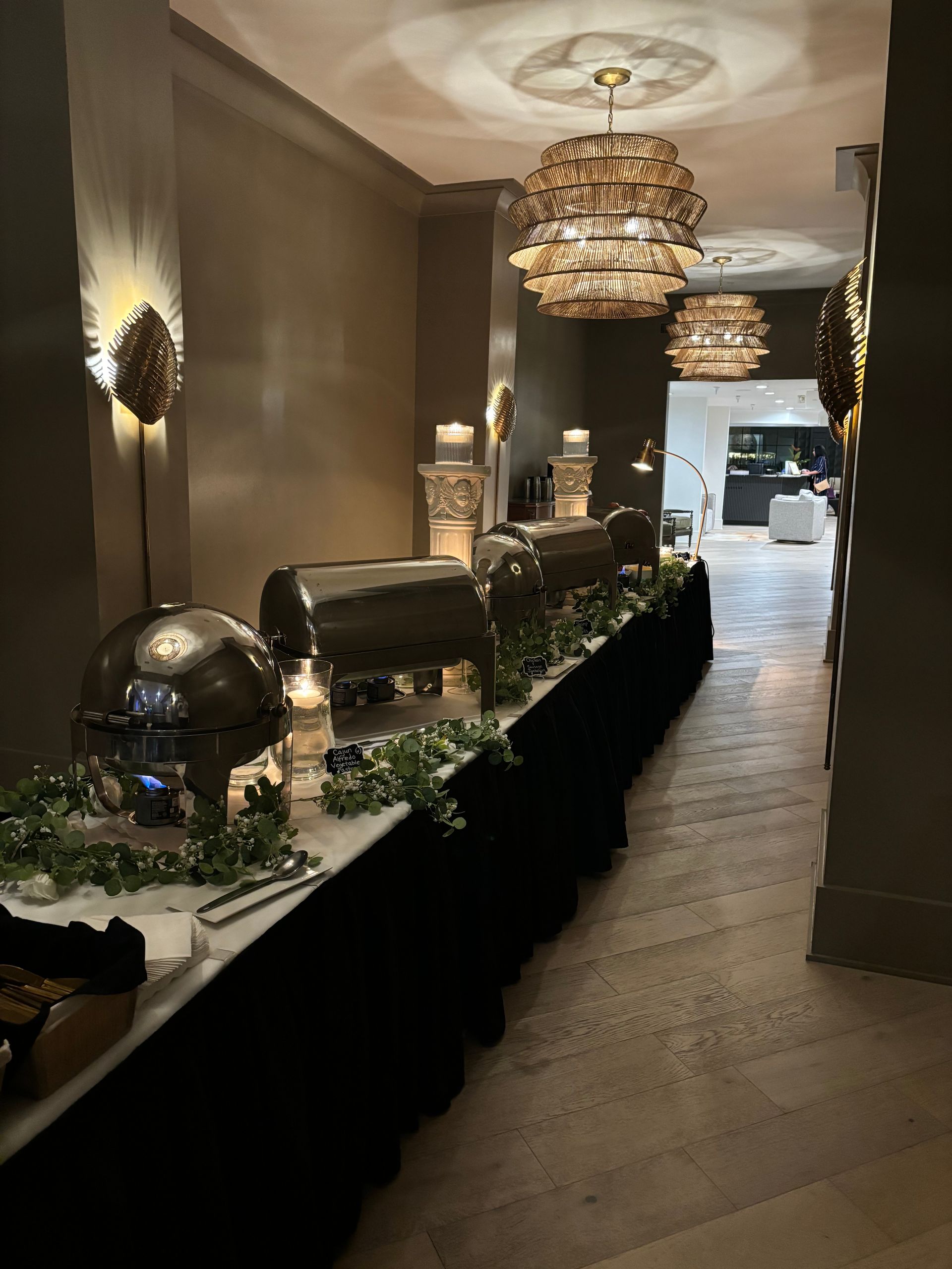 Buffet setup in a hallway. Silver food warmers on a long black-skirted table with greenery. Dim lighting and chandeliers.