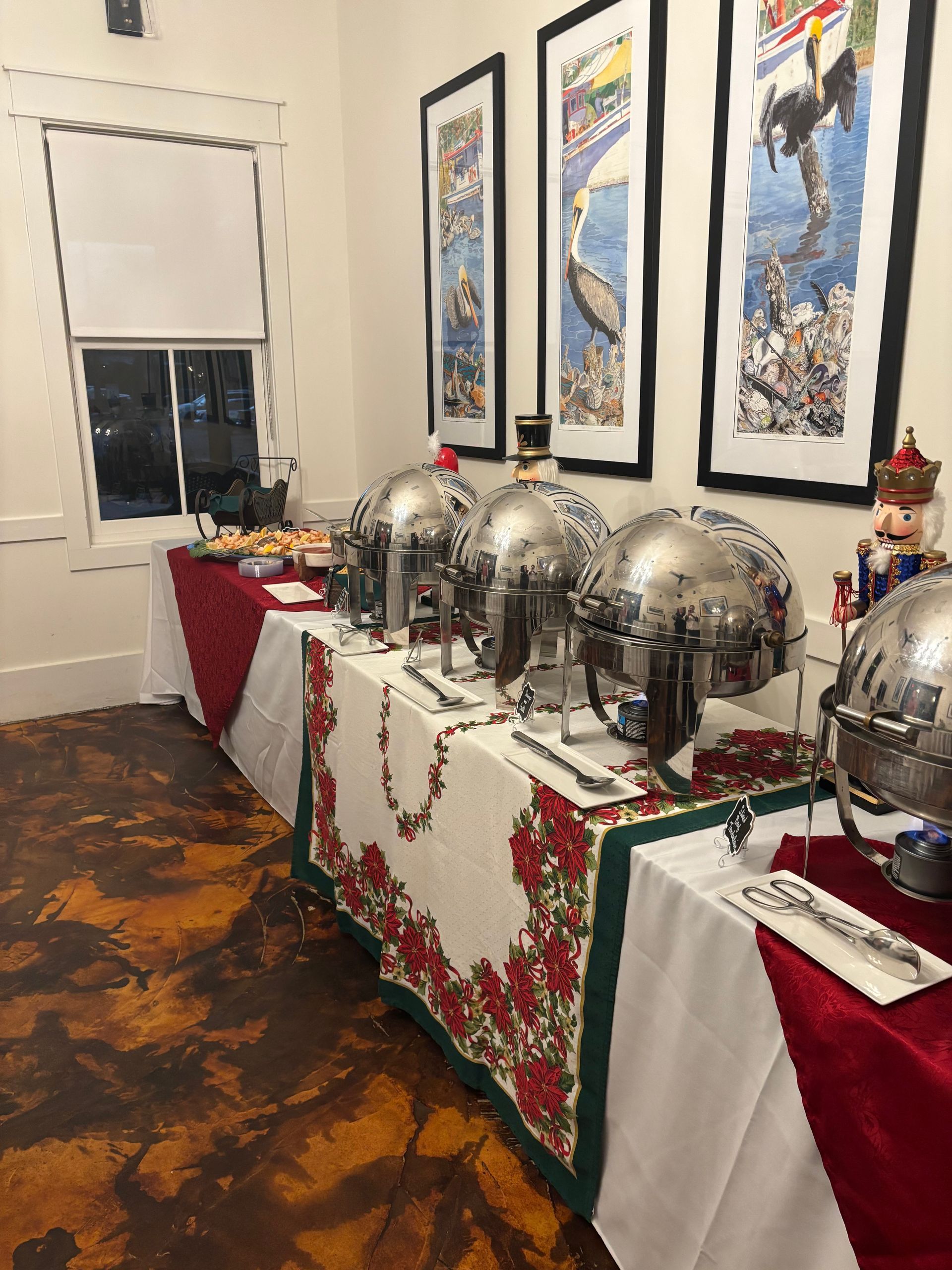 A buffet table is set up with silver chafing dishes and a red and white holiday tablecloth. Framed artwork hangs on the wall.