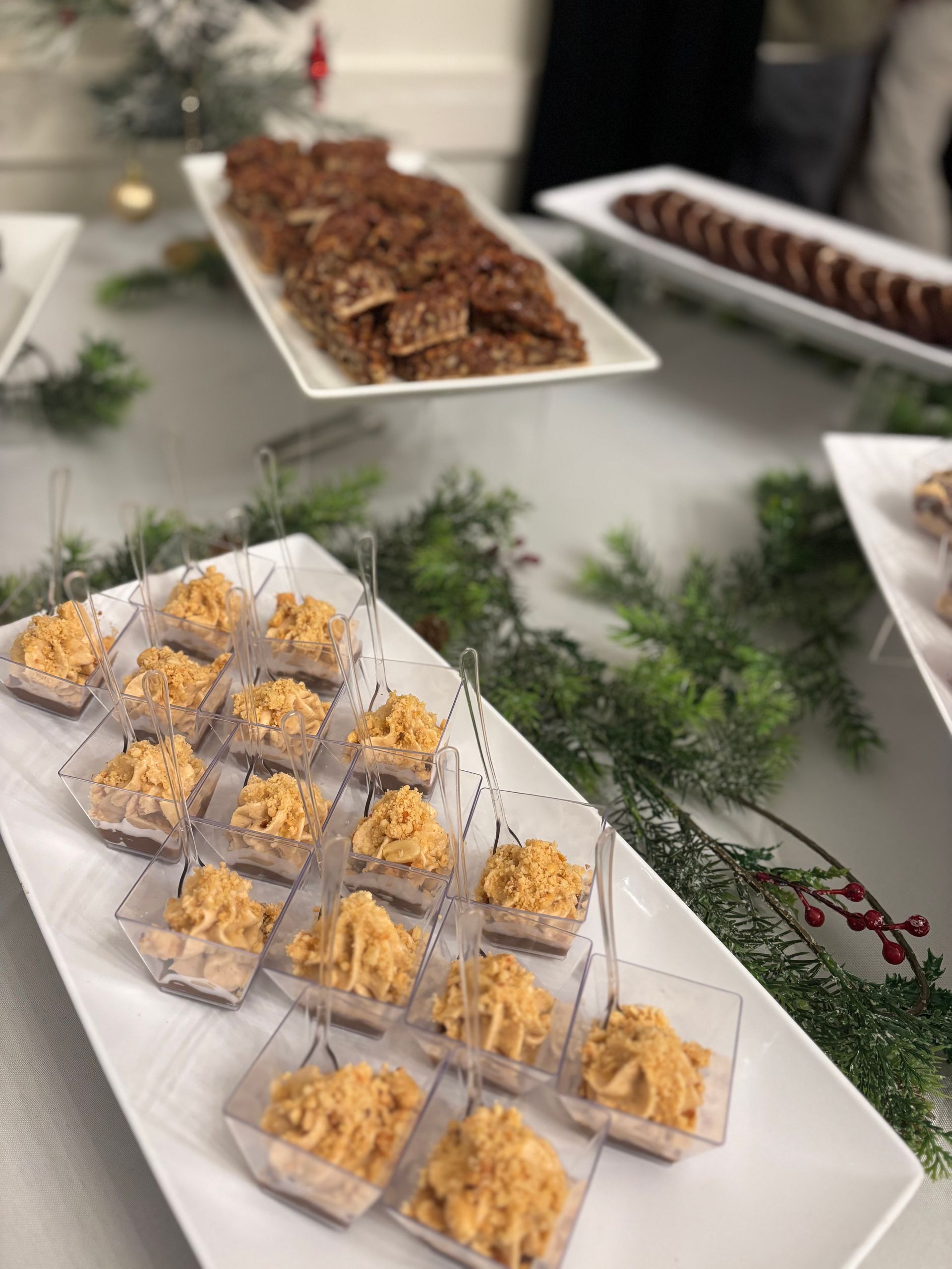A festive spread with appetizers: small cheese and cracker bites in clear cups, pecan brittle, and sliced sausage on white platters.