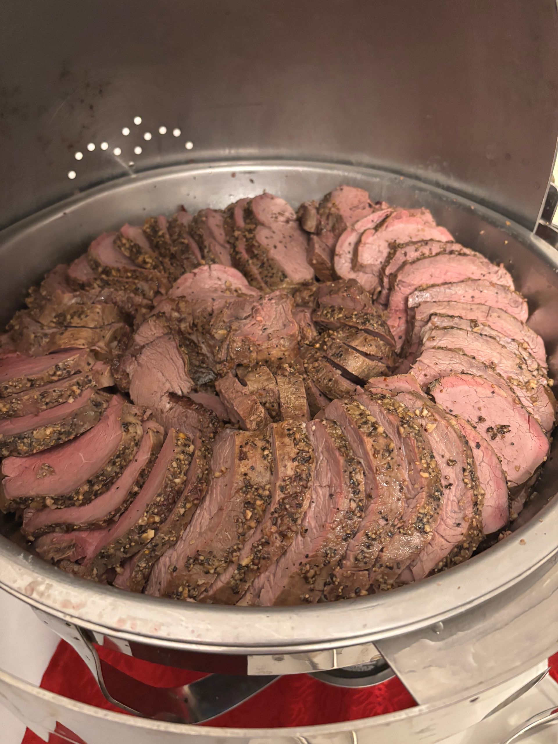 Sliced roast beef in a warming tray, arranged in a circular pattern. The beef is seasoned with herbs and spices and displayed at a buffet.