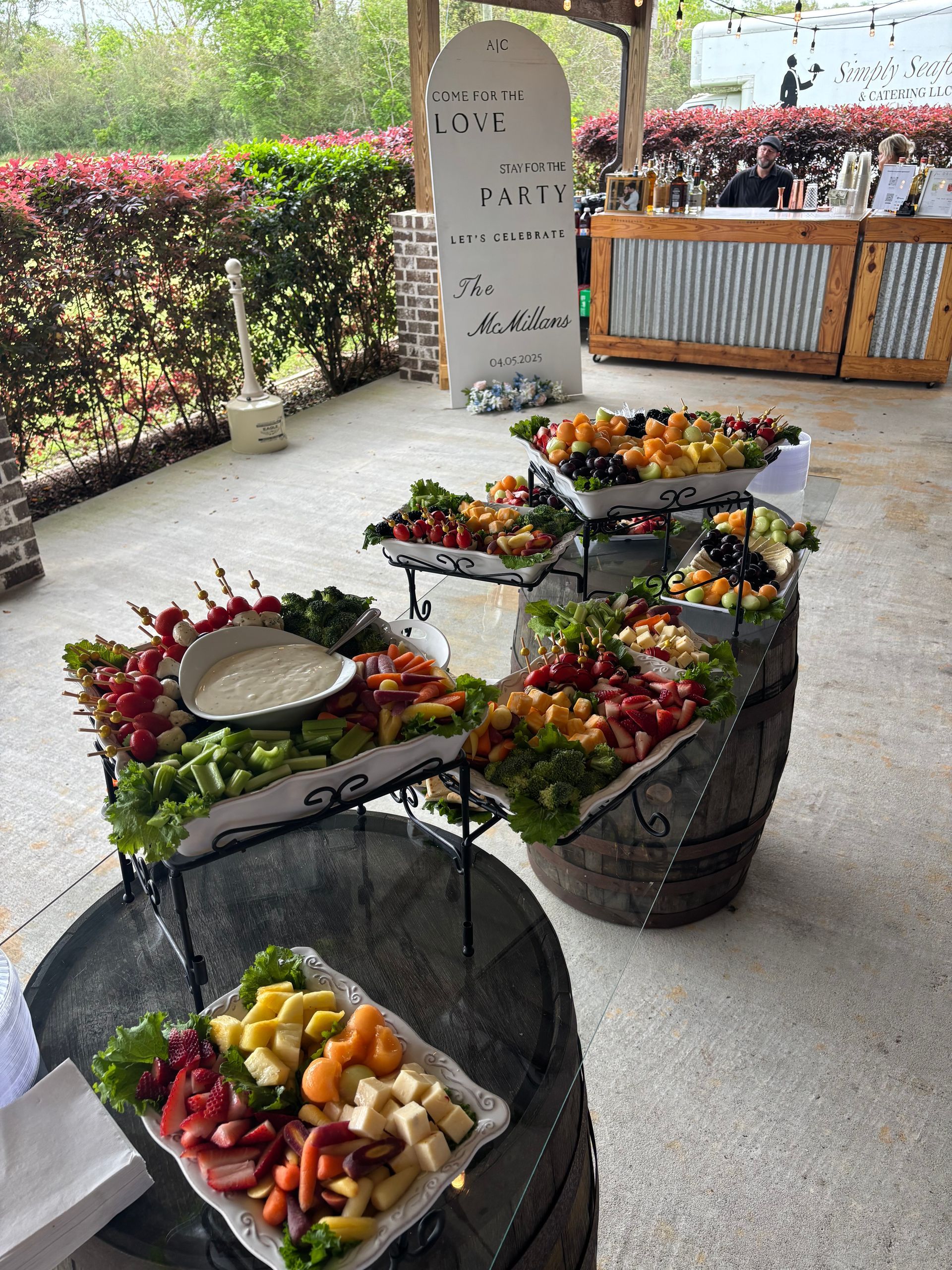 Outdoor buffet with platters of fruits and vegetables on barrels. A sign is visible in the background near a bar.
