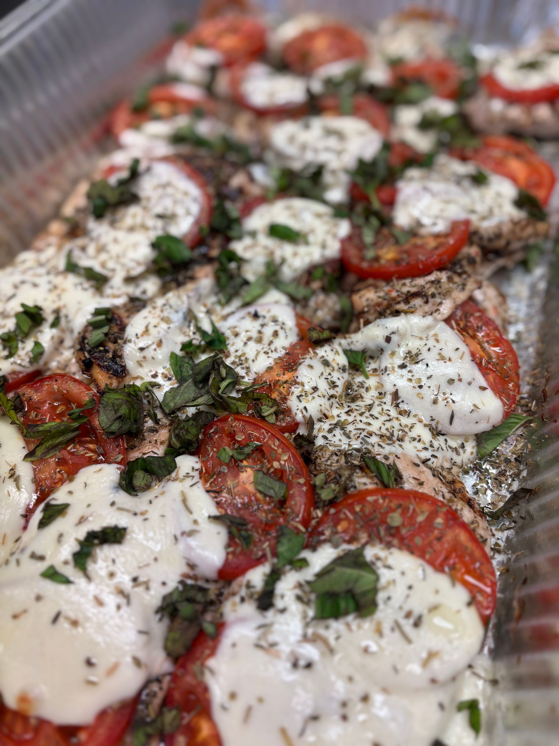 Baked dish of chicken breasts topped with sliced tomatoes, mozzarella cheese, basil, and herbs in a silver pan.