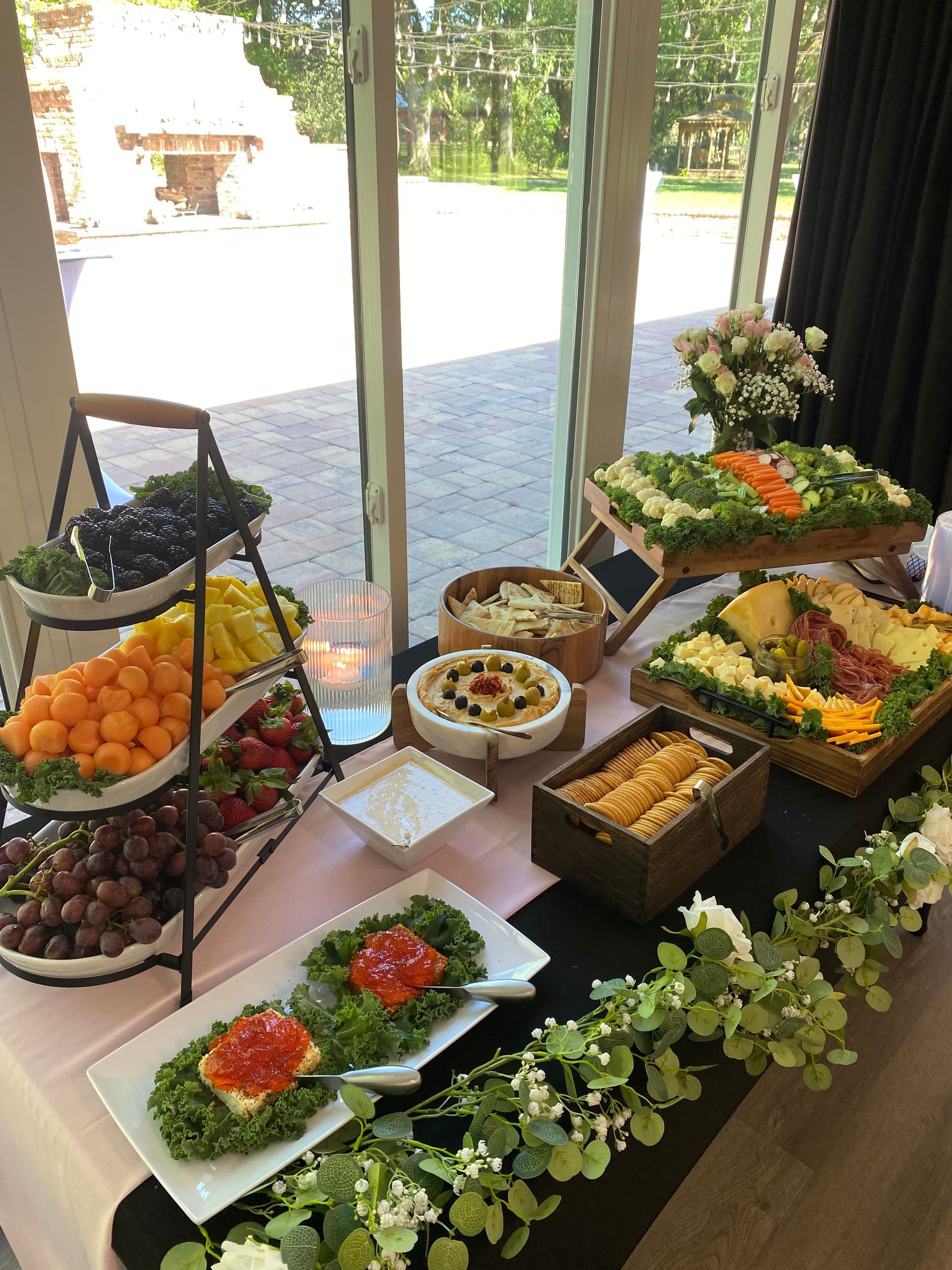 A table laden with a variety of food, including fruits, dips, and crackers, arranged near a large window.