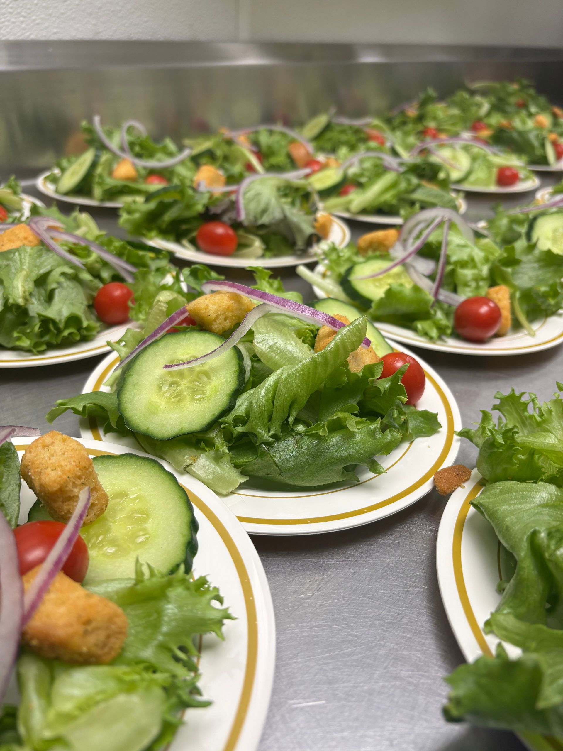 Salads on paper plates, featuring lettuce, cucumber, tomatoes, red onion, and croutons, prepared for serving.