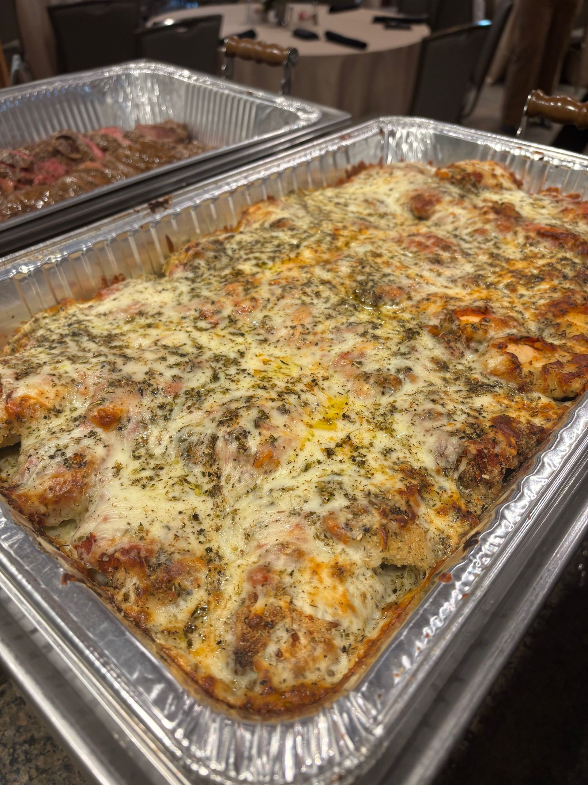 A metal tray overflowing with baked lasagna, topped with melted cheese and herbs. Another tray with food is visible in the background.