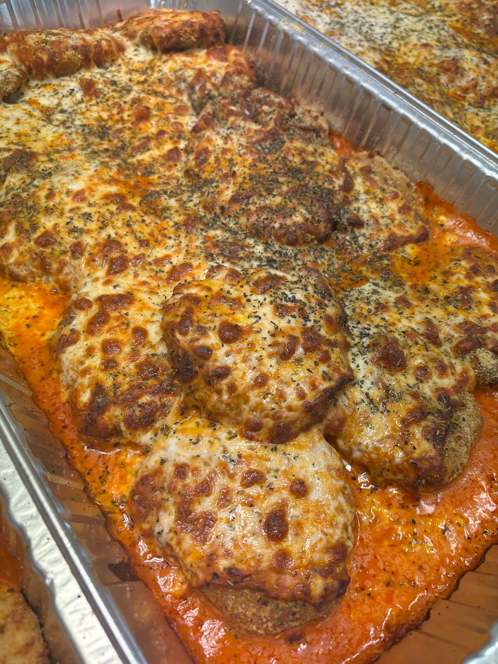 Tray of chicken parmesan, baked with melted cheese and herbs on a red sauce, in a metal pan.