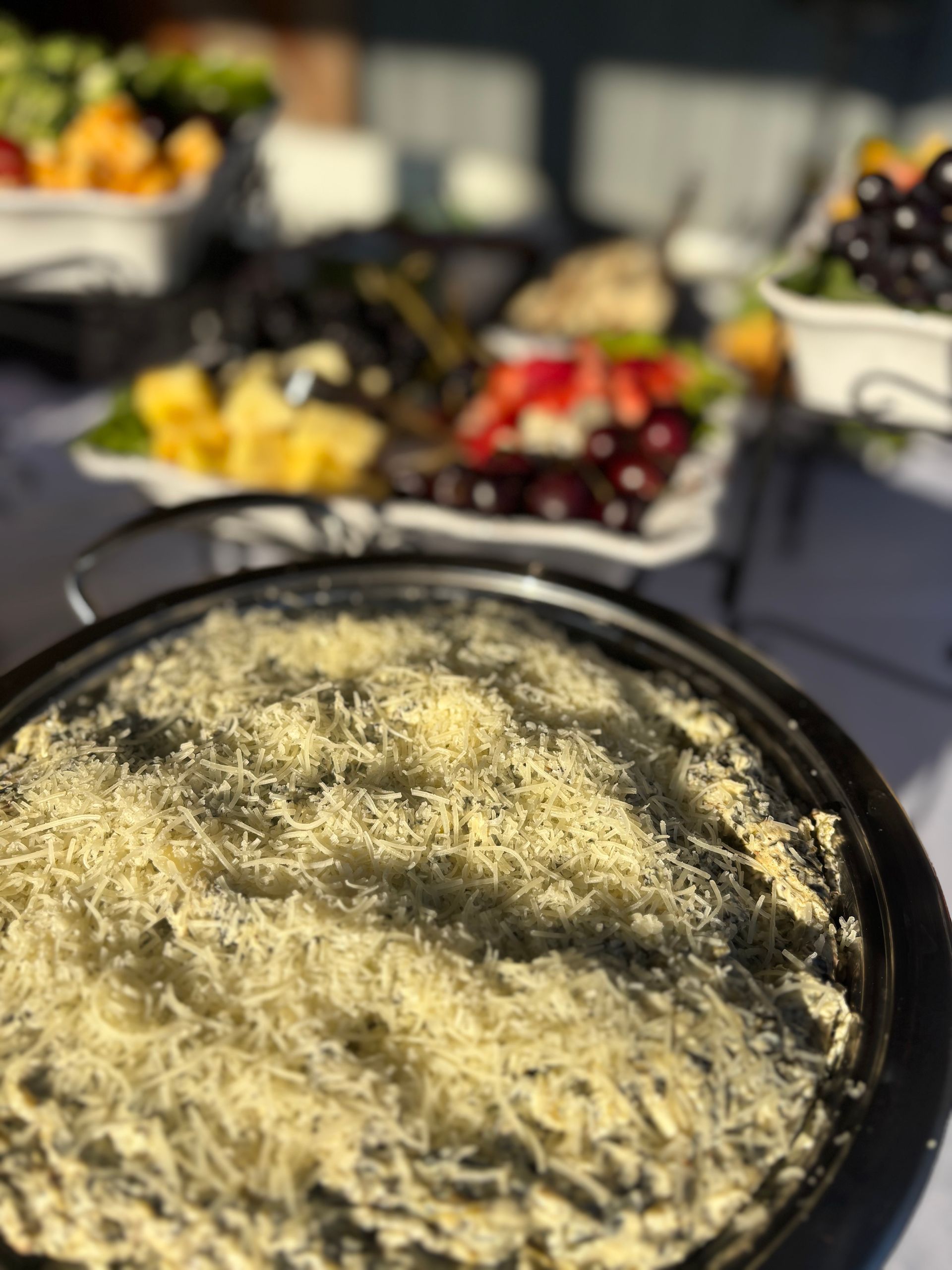 A close-up of a dish covered with shredded cheese, with a fruit platter in the blurred background.