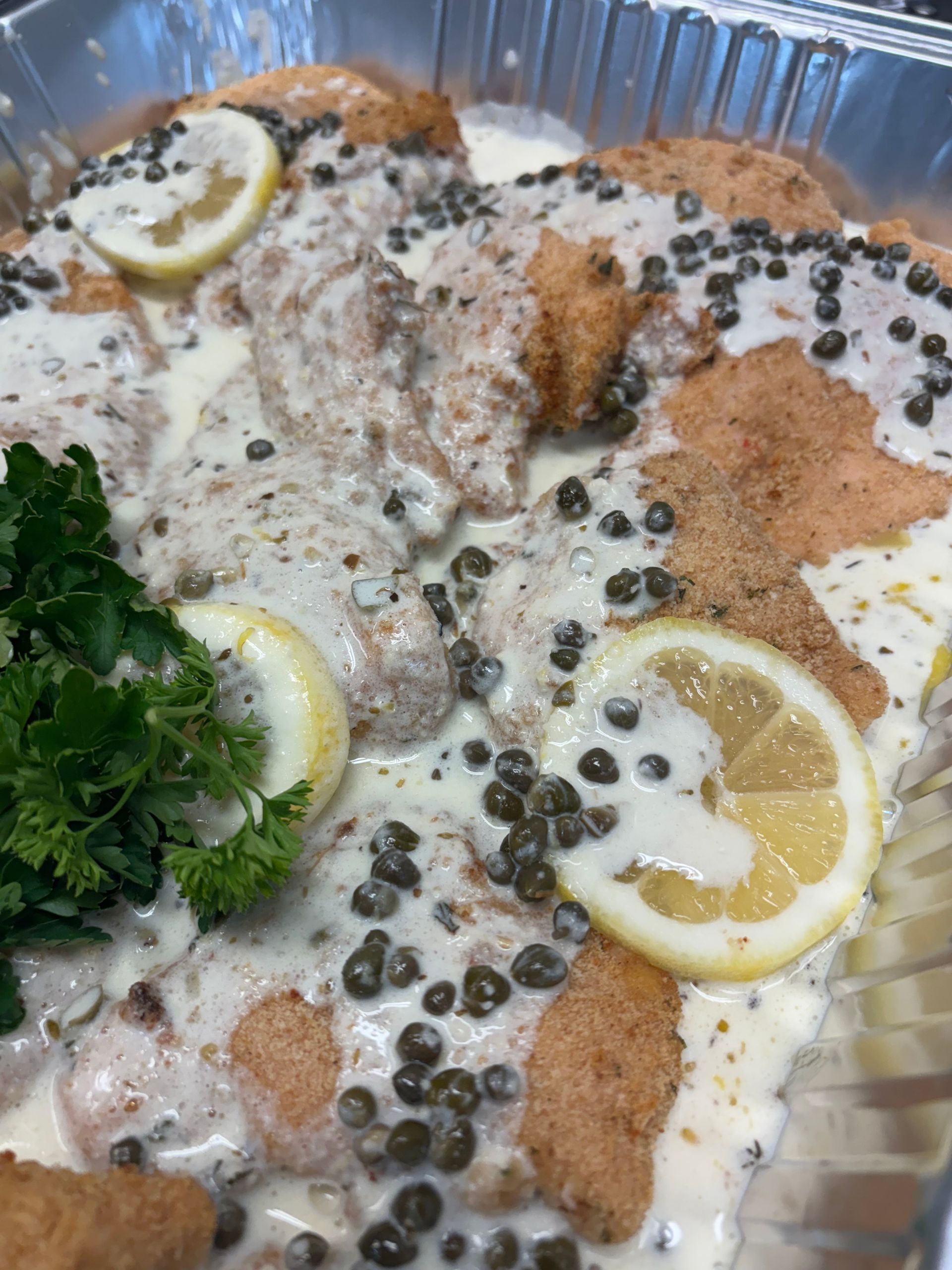 Chicken piccata in a foil pan, covered in creamy sauce, capers, lemon slices, and parsley.