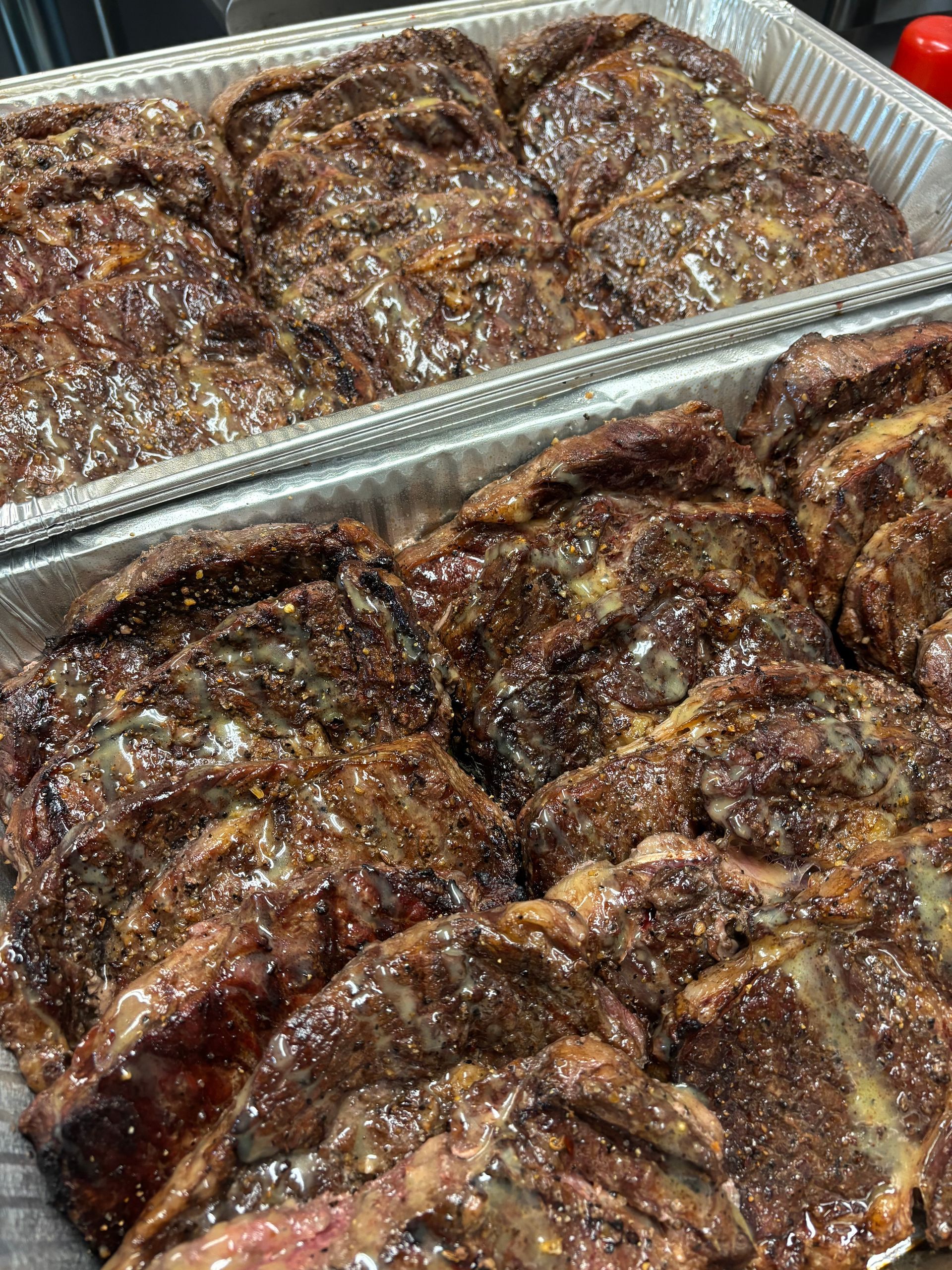 Close-up of trays filled with cooked, glazed ribs. The ribs are dark brown and glistening in an aluminum foil container.