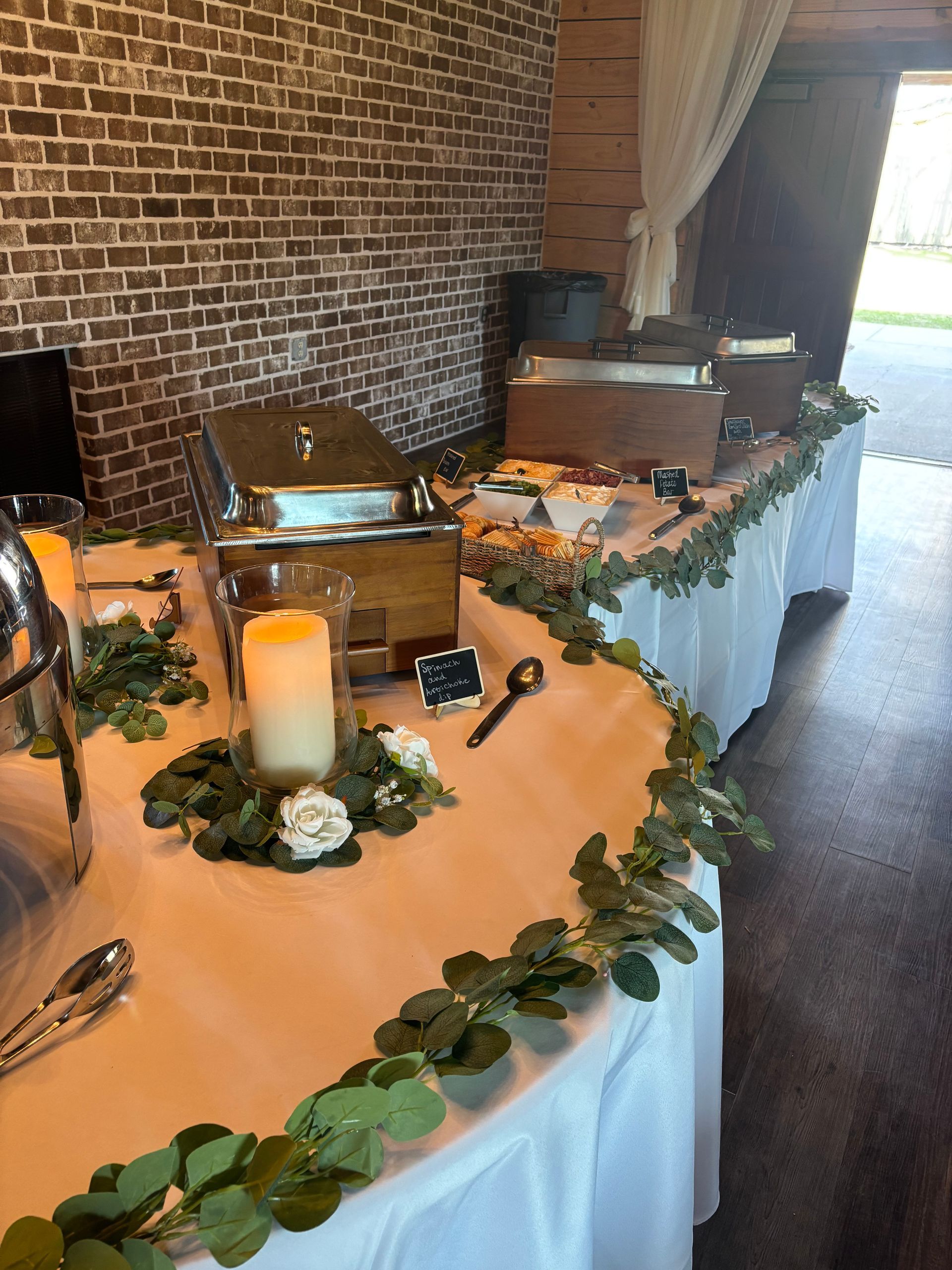 A buffet table with chafing dishes and food, decorated with greenery and candles, is set up inside a building with a brick wall background.