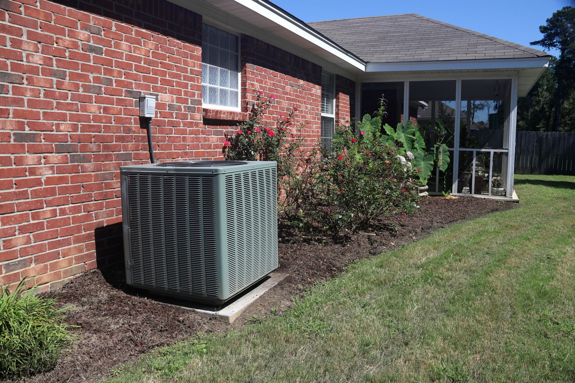 Air conditioner unit near a brick house with a screened porch, surrounded by mulch and greenery.