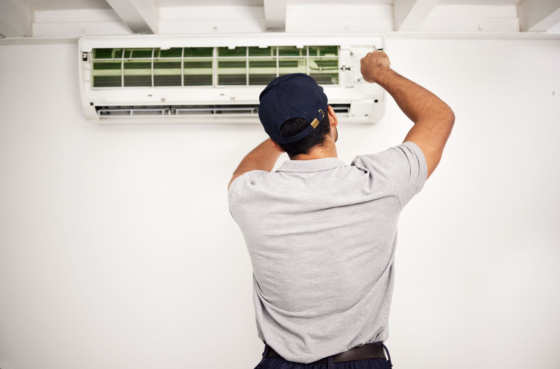 Man in a gray shirt and cap servicing a wall-mounted air conditioner indoors.