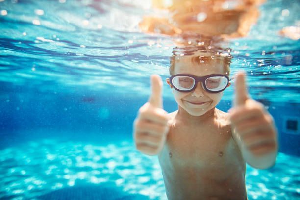 Boy with swim goggles underwater, giving thumbs up in a pool.