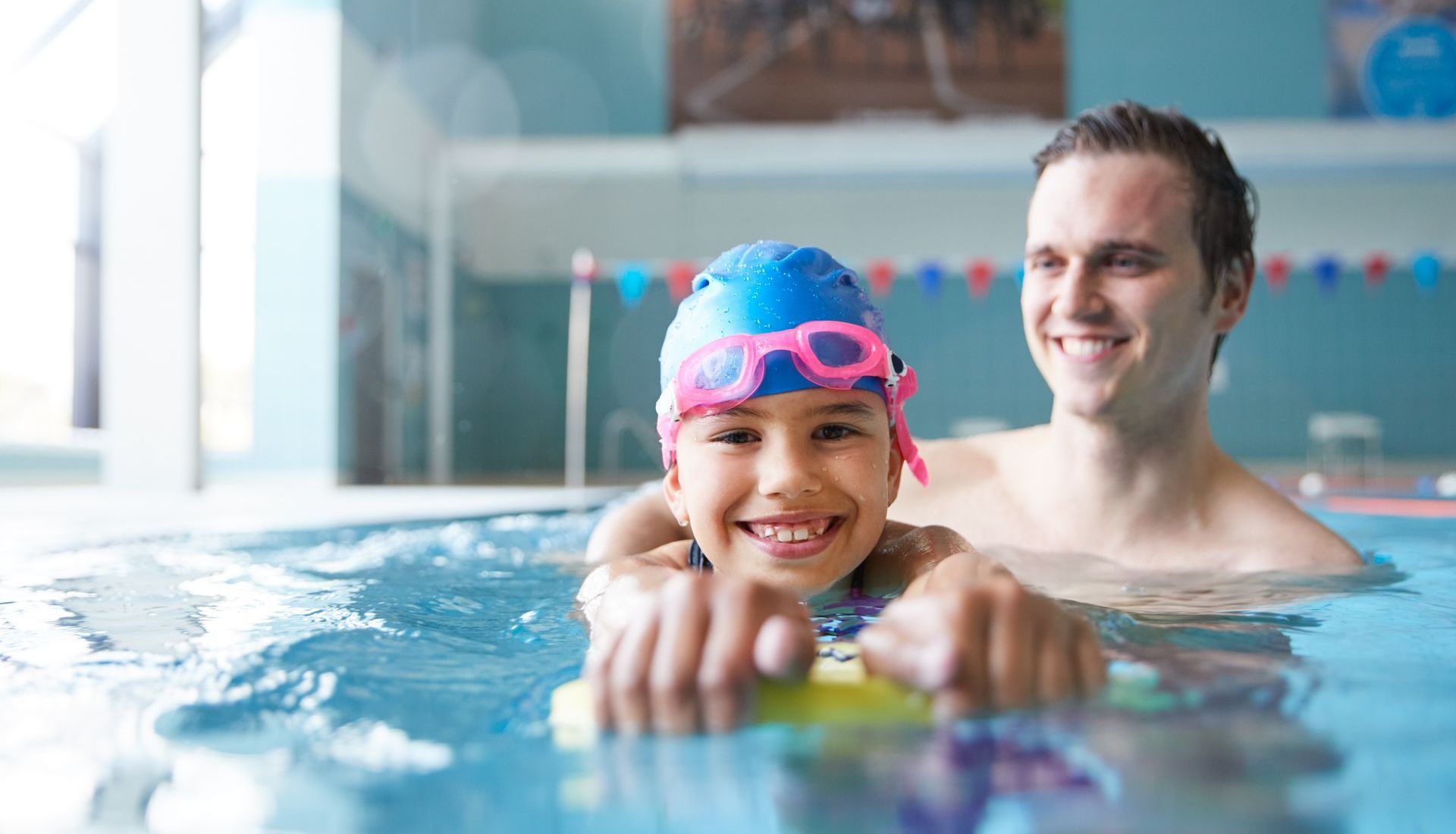 Child in blue swim cap and goggles smiles, learning to swim with an instructor in a pool.