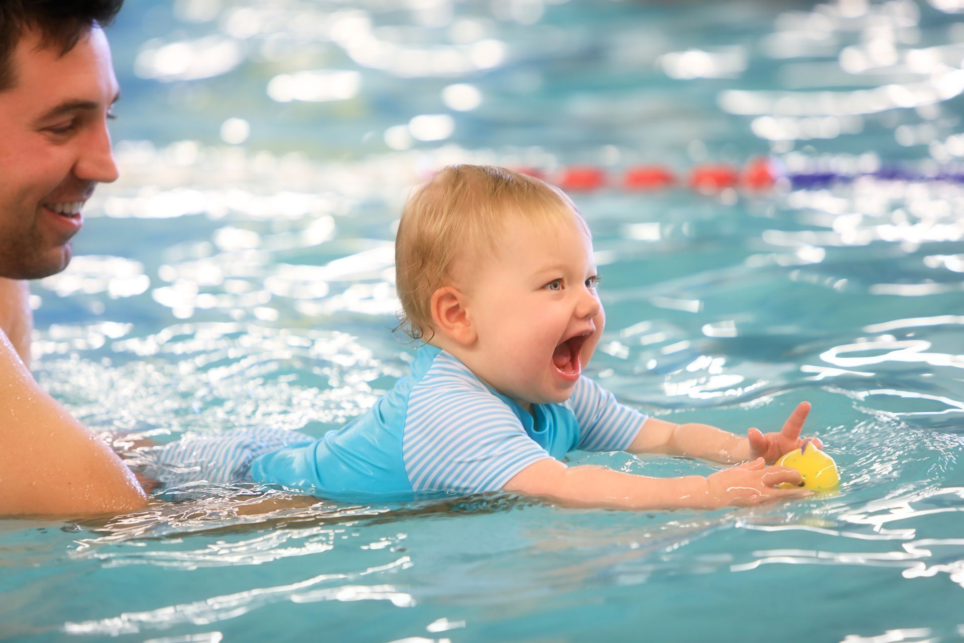 Smiling baby in blue swim shirt, floating in pool, with a smiling adult.