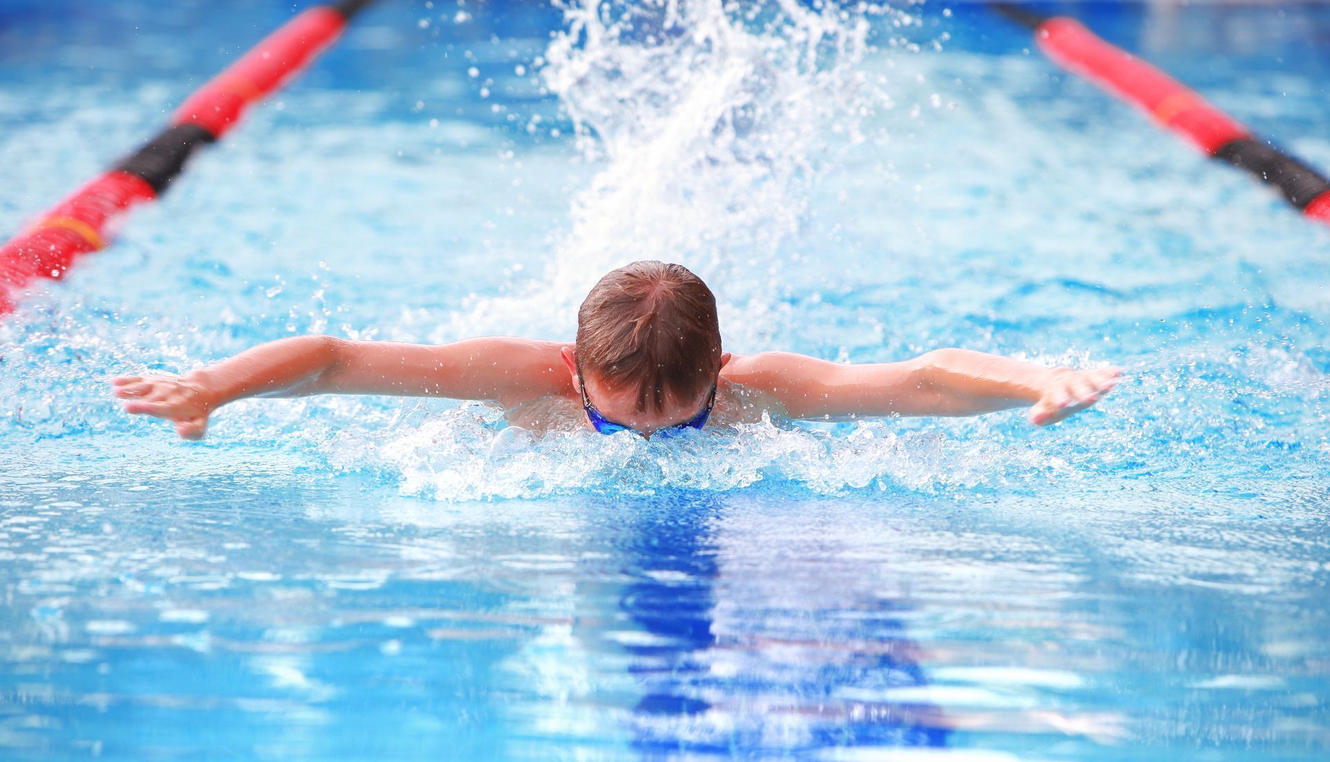 Young swimmer in blue goggles doing the butterfly stroke in a pool.