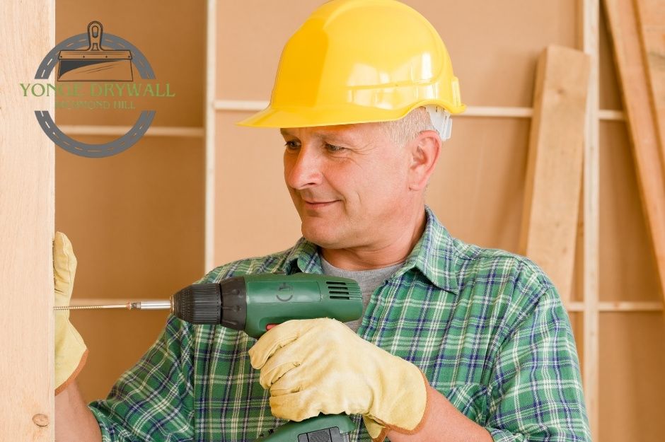 A drywall repair company worker wearing a yellow hard hat, green plaid shirt, and yellow gloves is using a green power drill to secure a screw into a wooden frame. The background shows unfinished drywall and wooden beams, indicating an indoor commercial construction site near King St, and Main St, Caledon, Ontario.