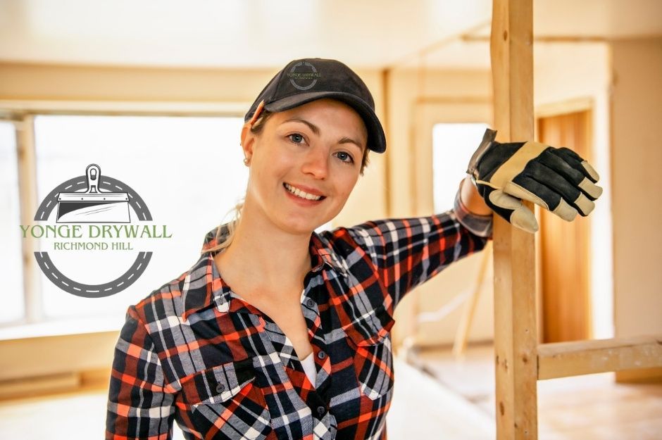 A drywall repair company employee smiling while wearing a plaid shirt, cap, and protective gloves. She is leaning against a wooden frame inside a bright, unfinished room in a commercial construction site near Concession Rd 4, and Squair Rd, Clarington, ON.