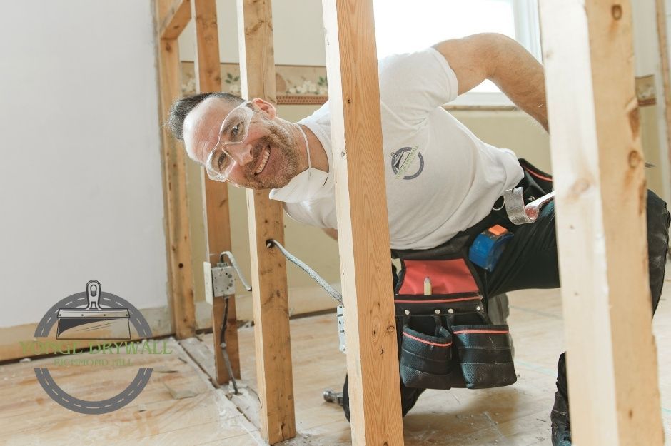 A drywall repair company worker wearing safety goggles, a white t-shirt, and a dust mask kneels between wooden wall studs during a residential renovation. He smiles while holding a pry bar and wears a tool belt with repair tools attached. Located near Banner Ln, and Elizabeth Grove, King, ON.